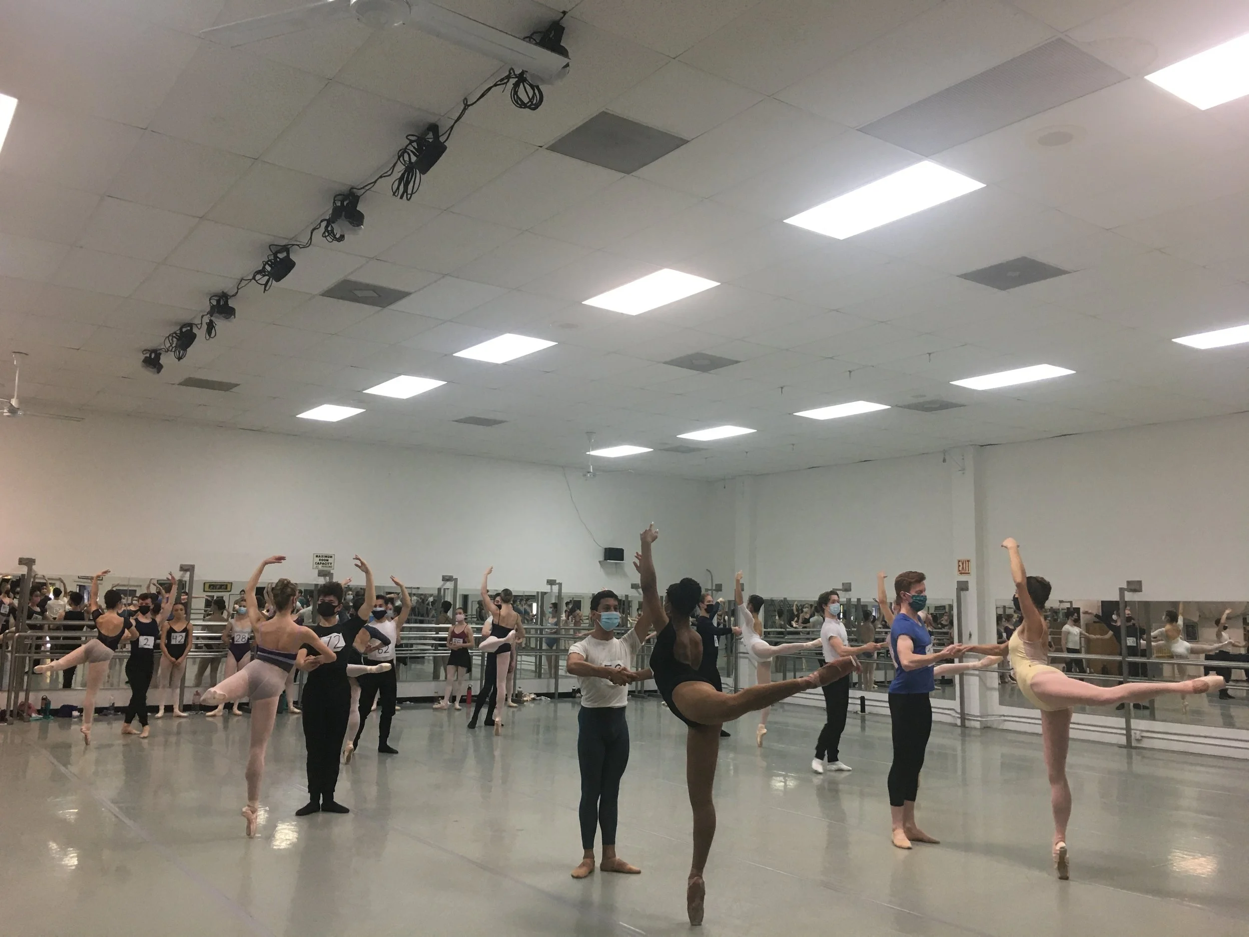 Ballet dancers in a dance studio practicing ballet, some holding poses and wearing masks.