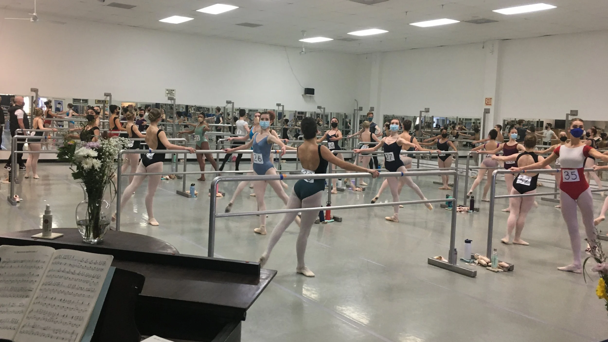 Ballet dancers practicing at a barre in a dance studio with many mirror panels, most of the dancers wearing face masks.