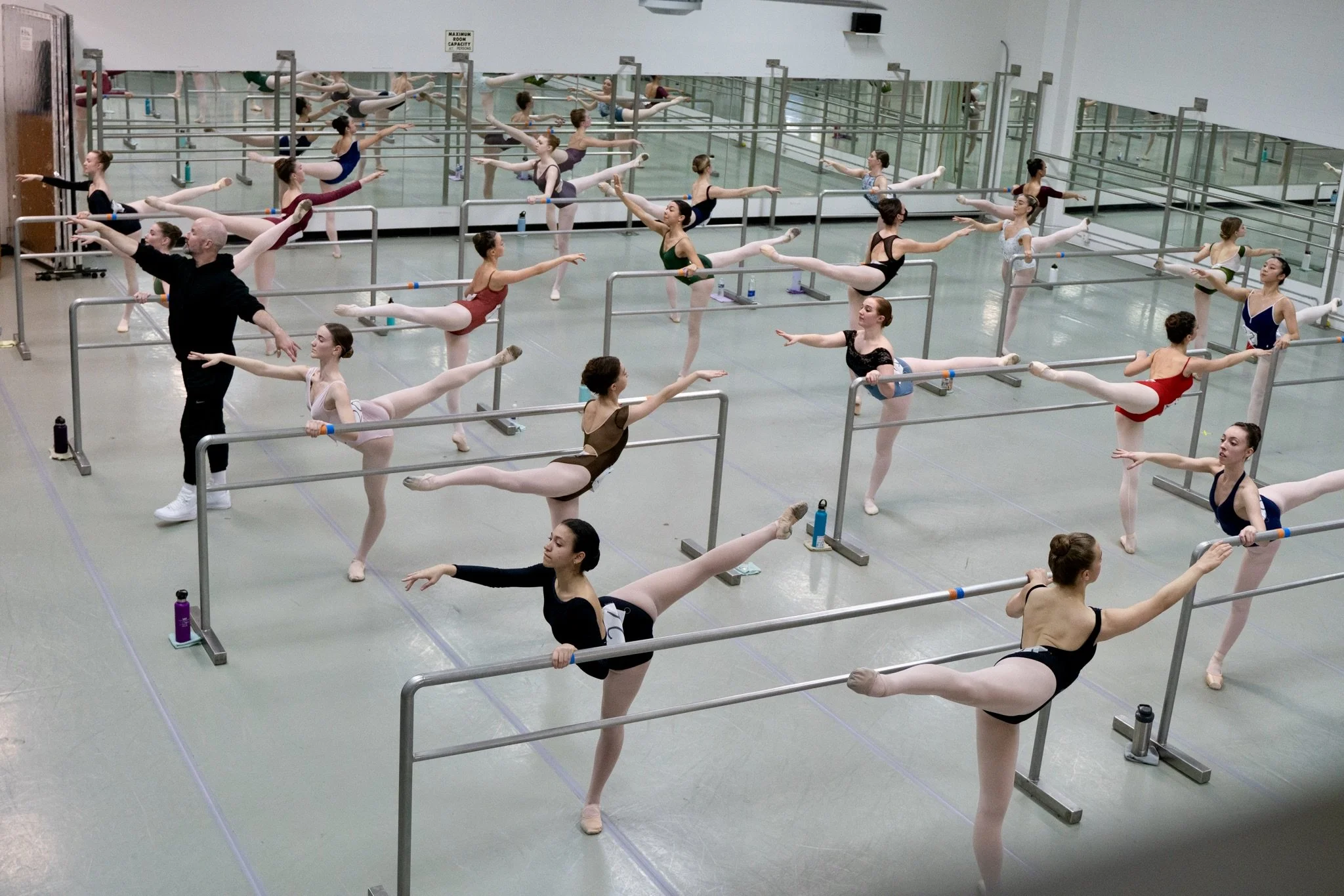 Ballet dancers in a dance studio practicing at the barre, with an instructor supervising. The studio has mirrors along the wall, and the dancers are stretching and warming up.