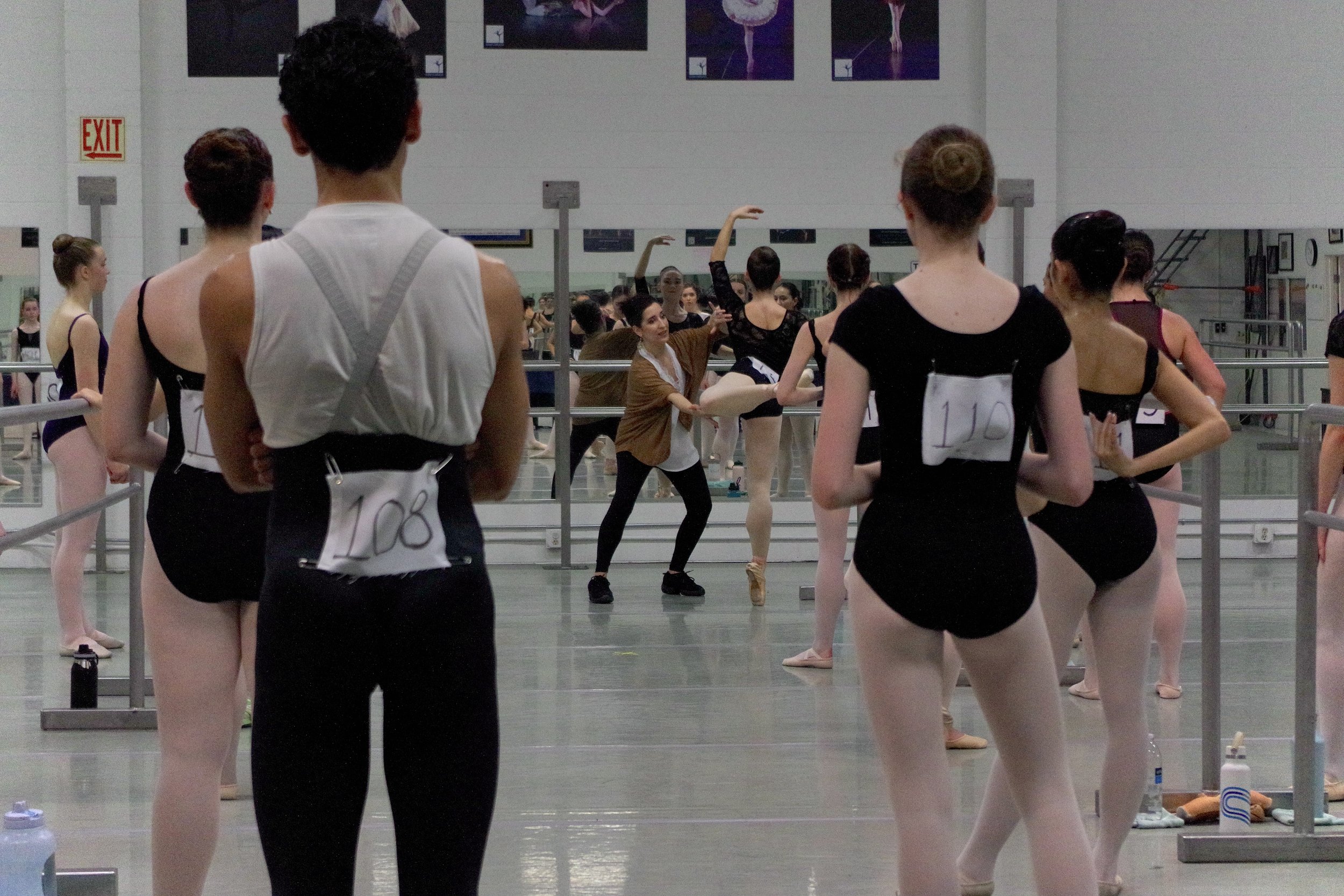 Dance class with students in black leotards and tights, instructor demonstrating dance moves in the center, mirrors and ballet barres in the background.