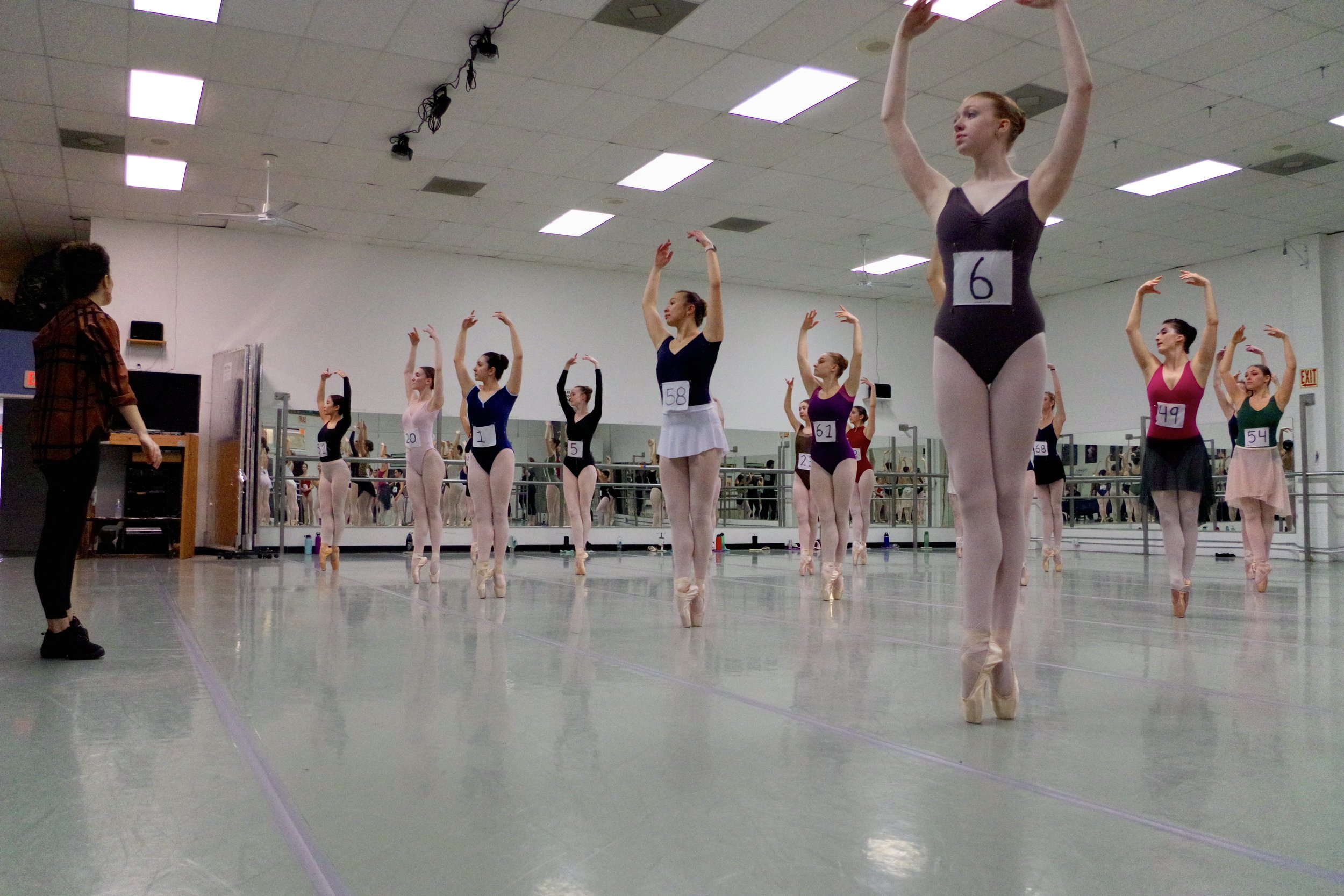 A group of ballerinas practicing at a dance studio with a teacher observing, all wearing numbered costumes and pointe shoes.