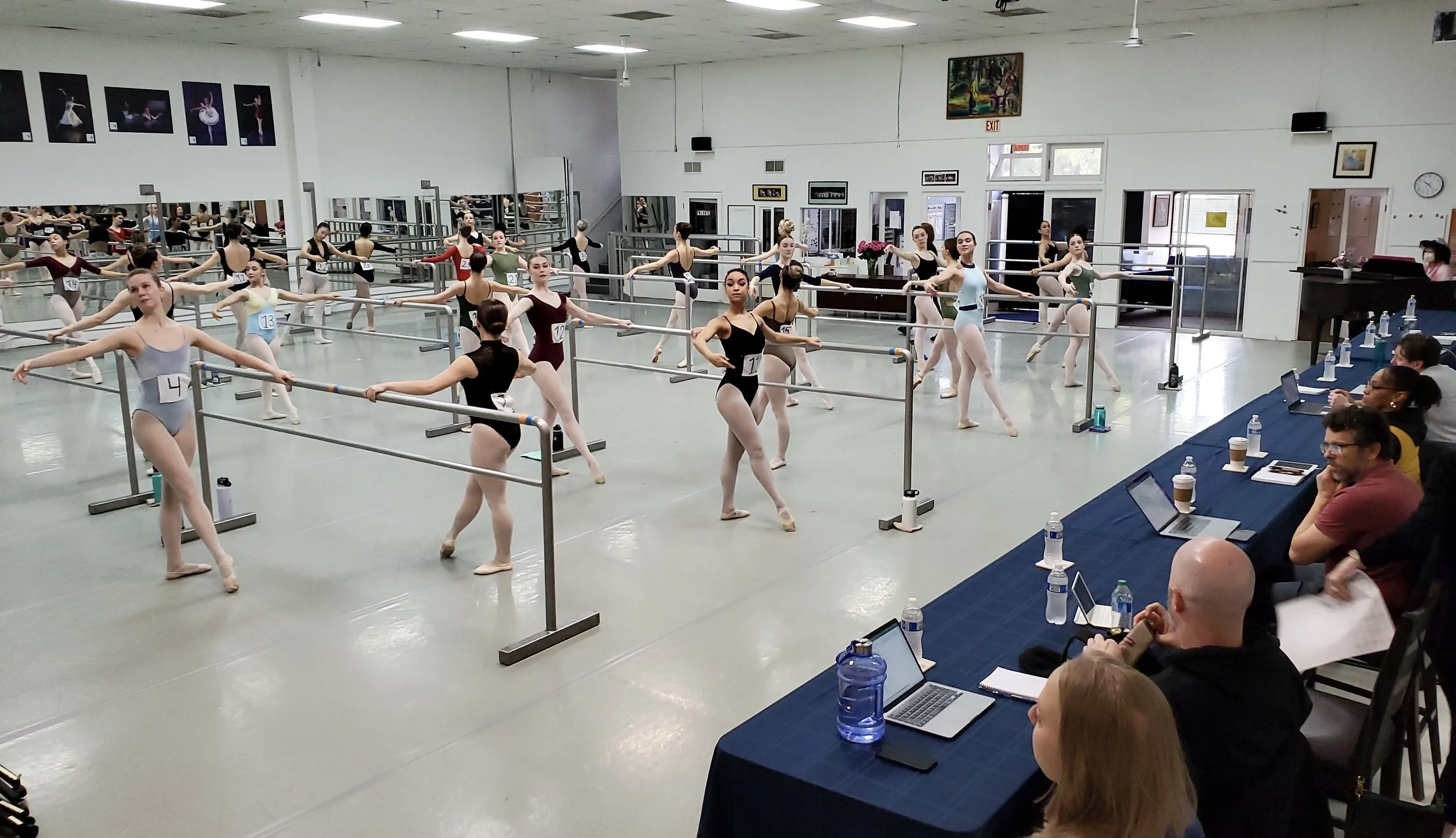 Ballet students practicing at a dance studio with judges observing from a table on the side.