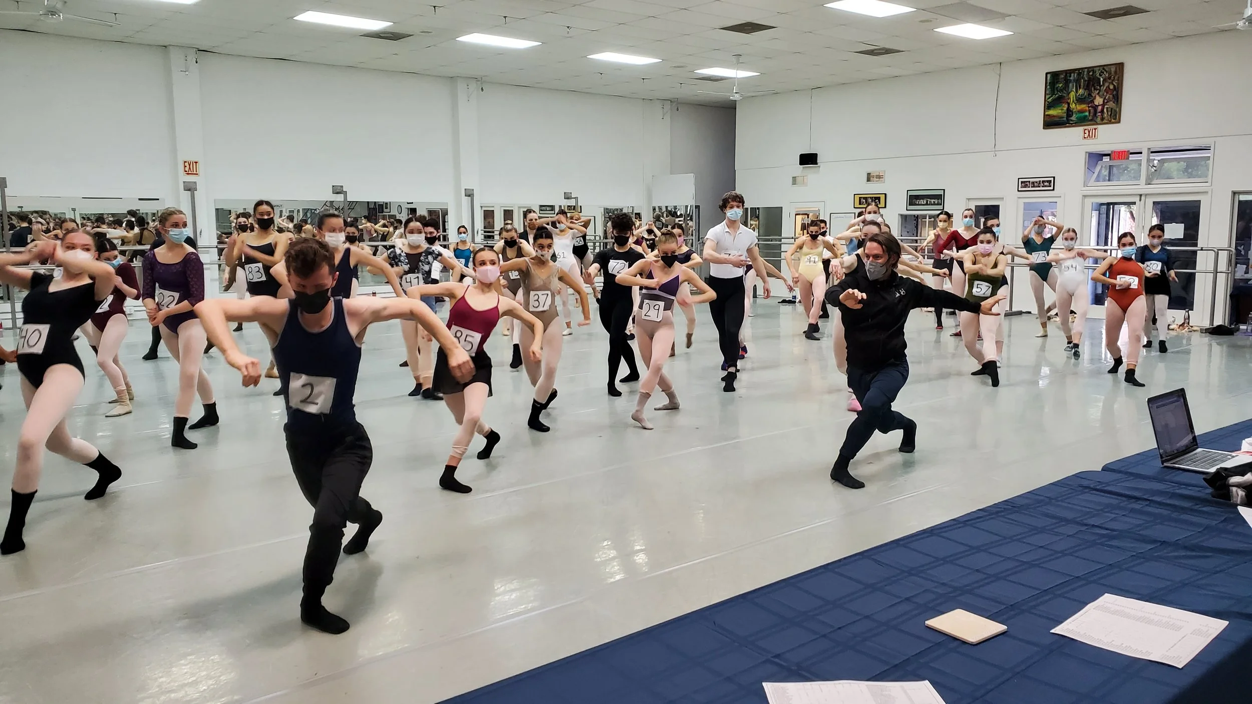 Ballet dancers practicing in a studio, wearing masks and numbered costumes, instructor leading the session, masks on all individuals, tables with papers and laptop in foreground.
