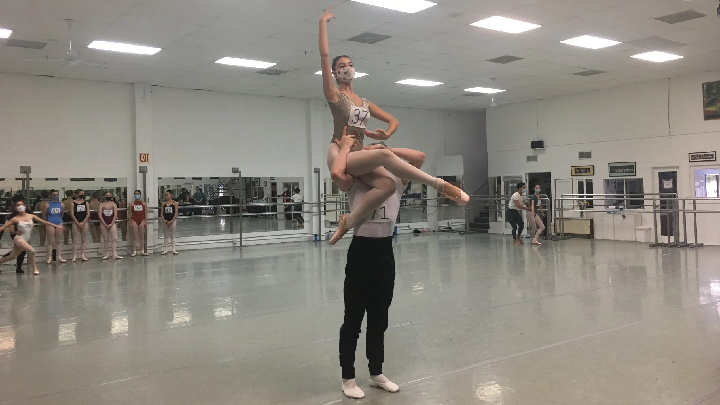 Ballet dancers practicing in a dance studio, one being lifted by another, with other dancers observing in the background.