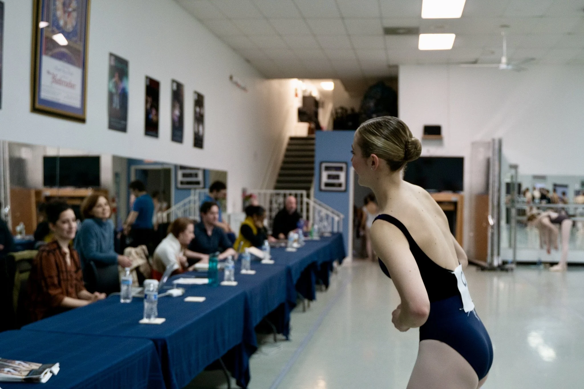A young female gymnast in a black leotard preparing for her performance in a gymnasium with judges seated at a long table in front of her.