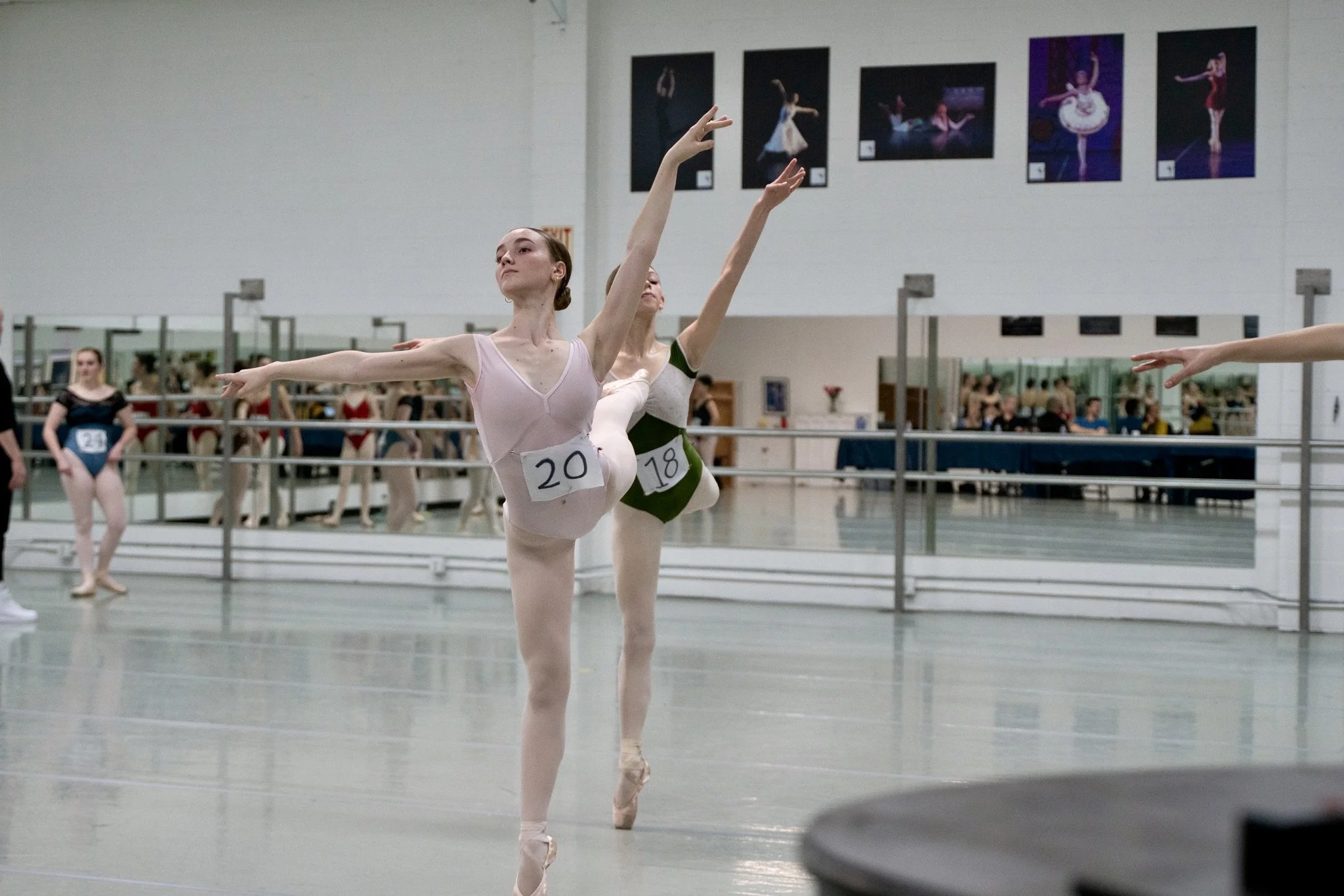 Ballet dancers practicing in a dance studio, two dancers in the foreground performing pirouettes, with others visible in the background and on wall posters.