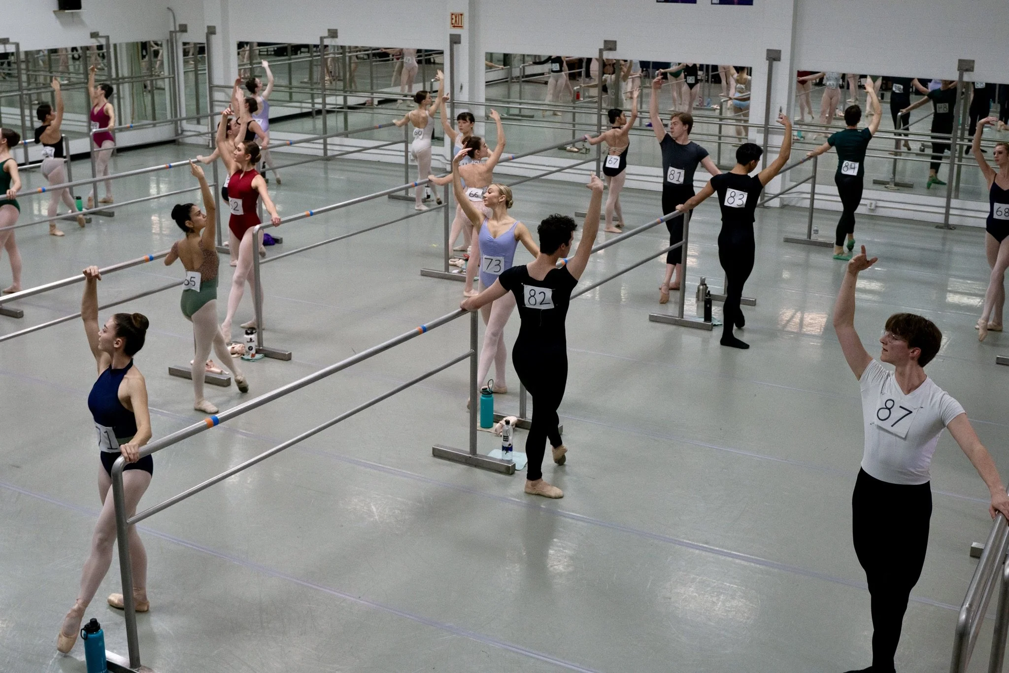 Ballet dancers in a dance studio practicing at ballet barres, with some stretching and warming up, reflected in mirrored walls.