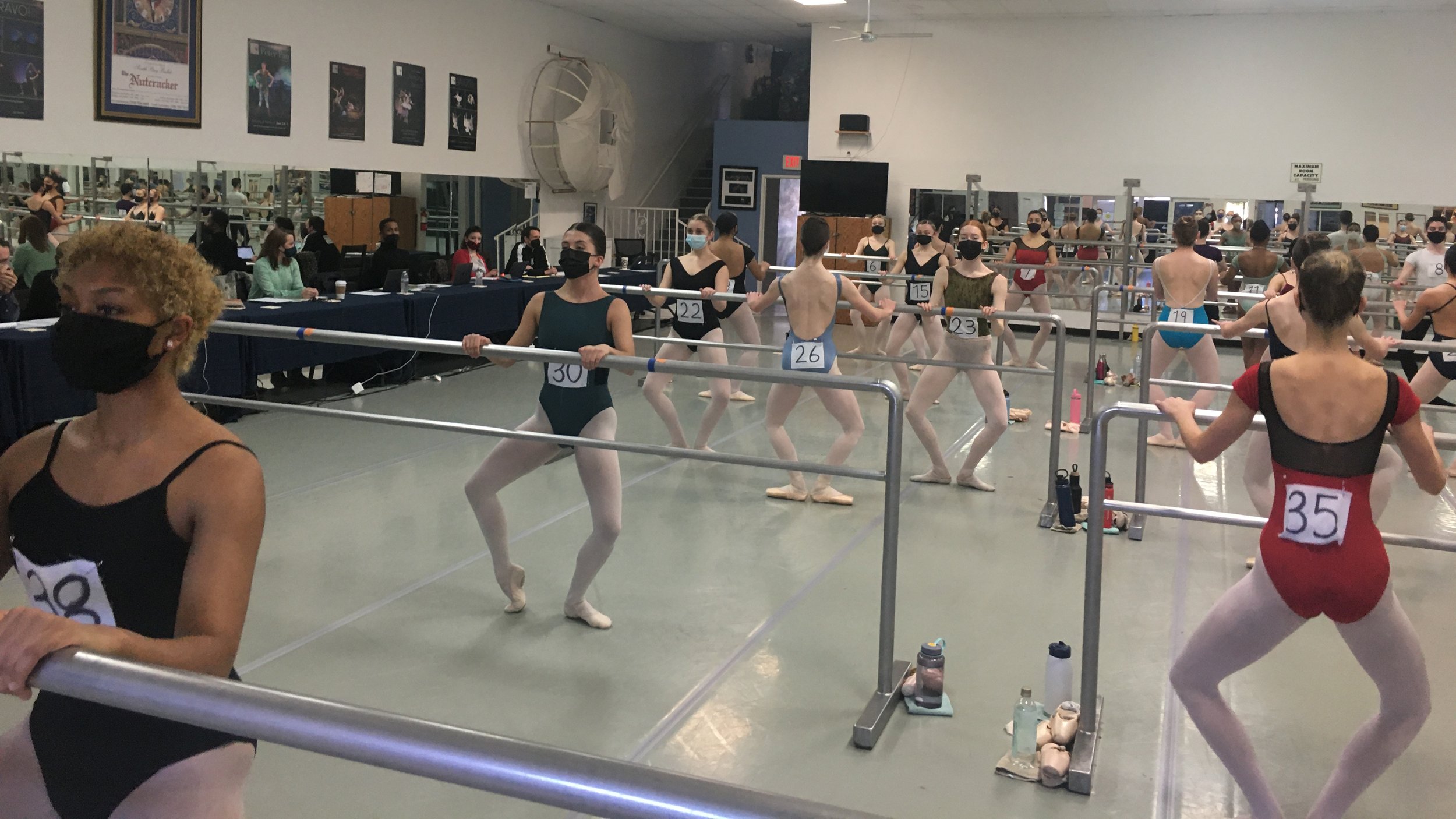 Ballet dancers practicing at a ballet school, with some wearing masks and numbered costumes, and others sitting at a table in the background.