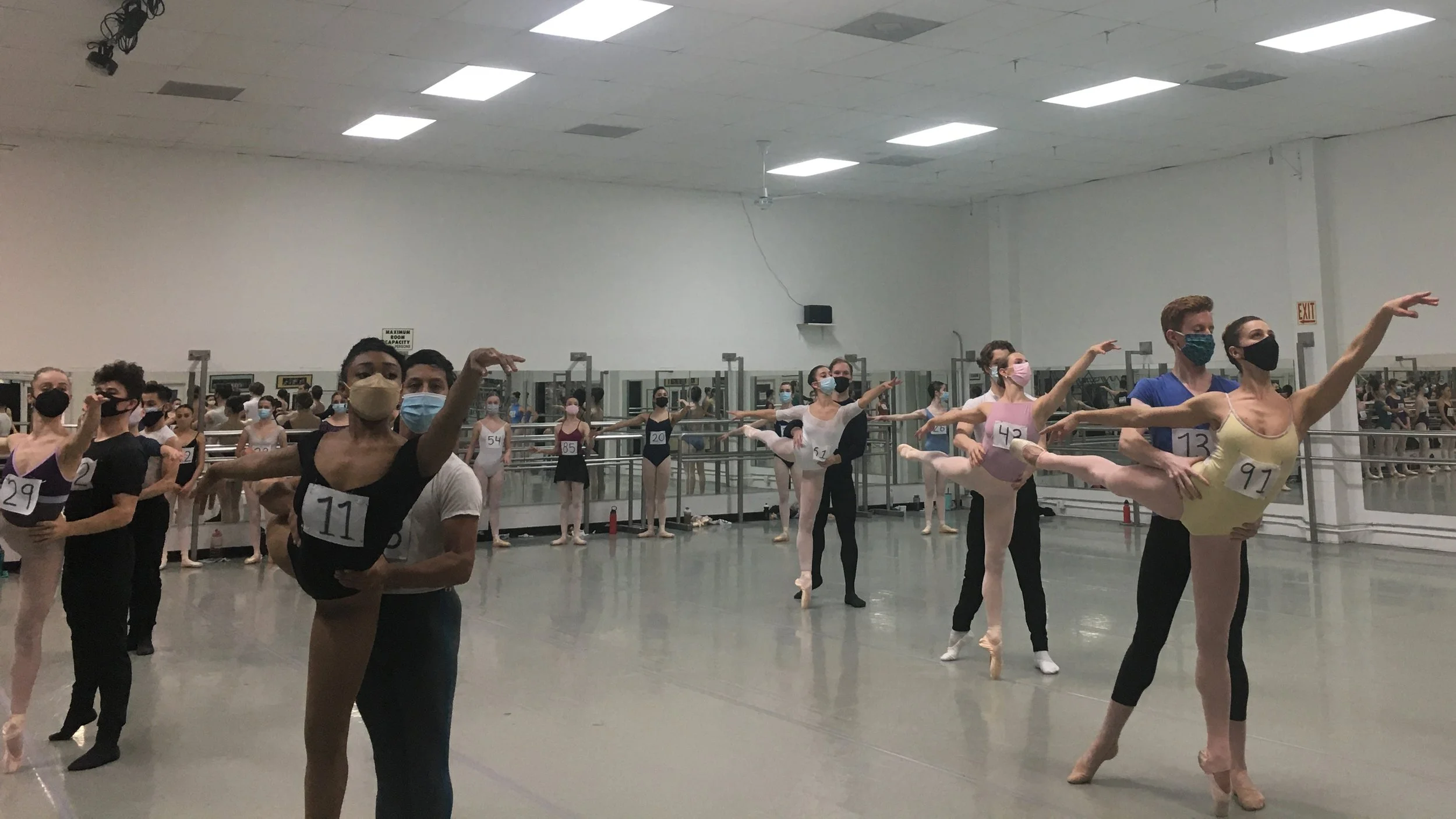 Ballet dancers practicing in a dance studio, all wearing masks and numbered costumes, with some holding poses.