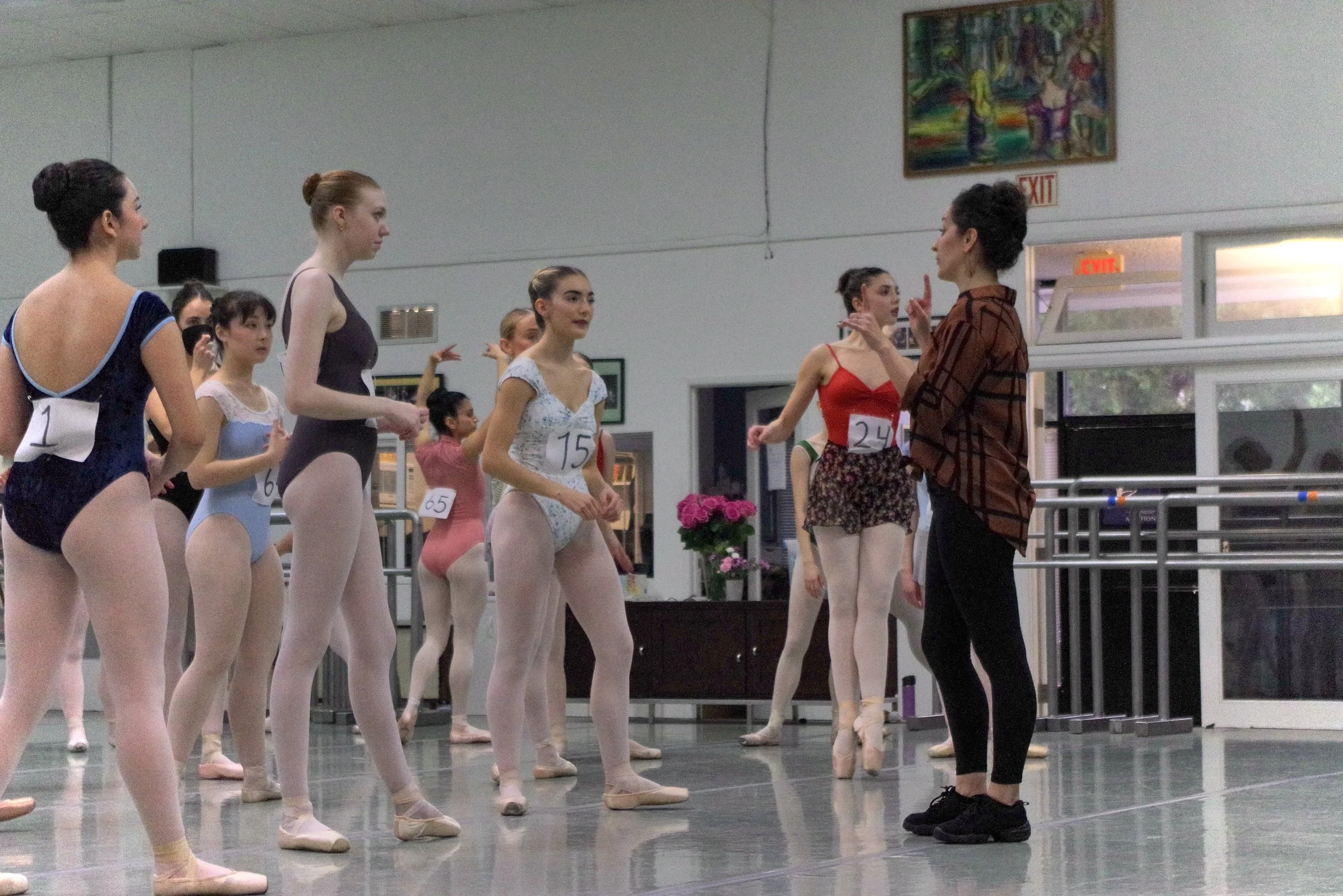 Ballet dancers in practice leotards and tights listening to their instructor, who is giving instructions inside a dance studio.