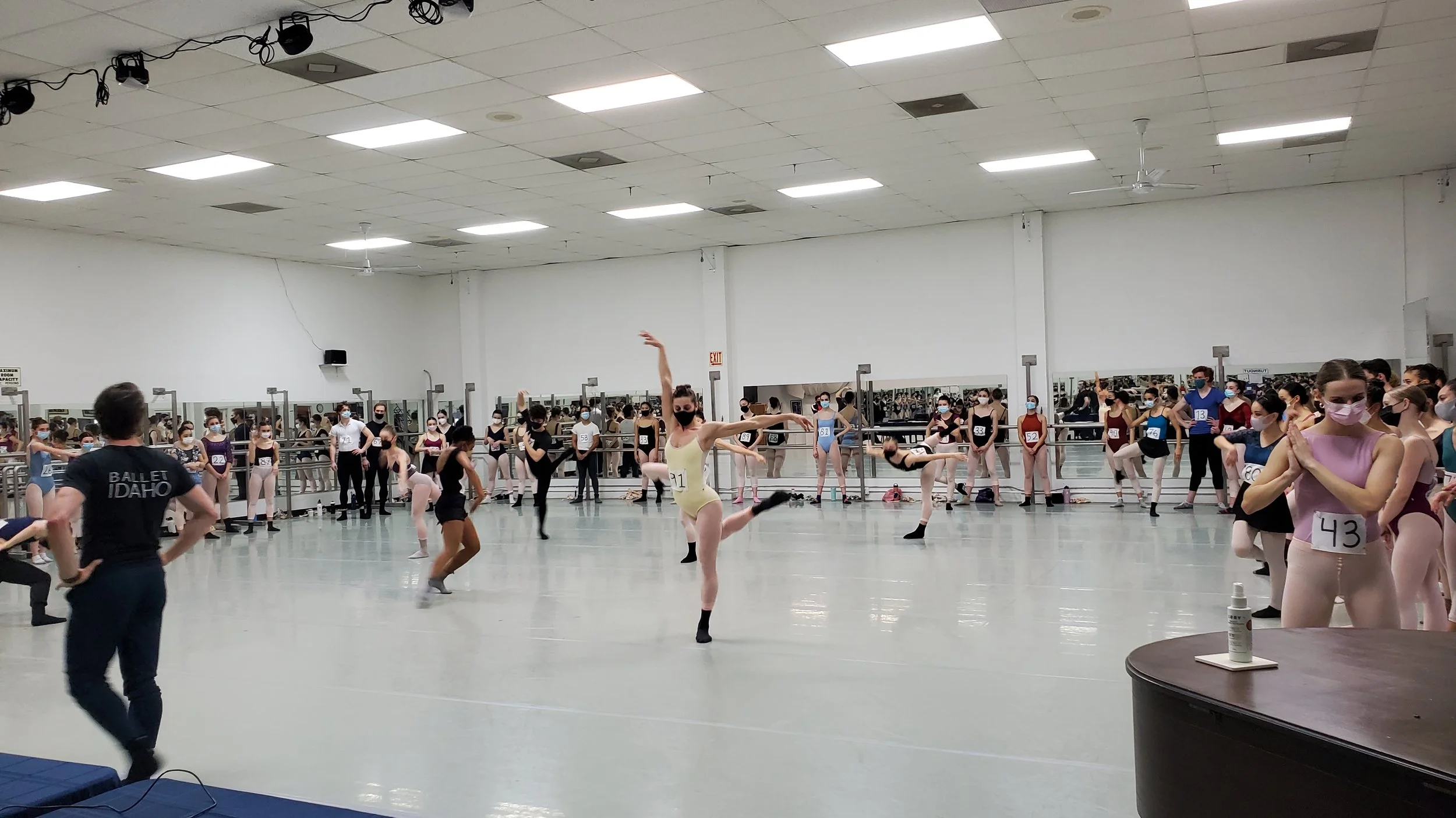 Ballet dancers practicing in a dance studio with mirrors, some dancers are stretching or warming up, all wearing masks, with a person observing in the foreground.