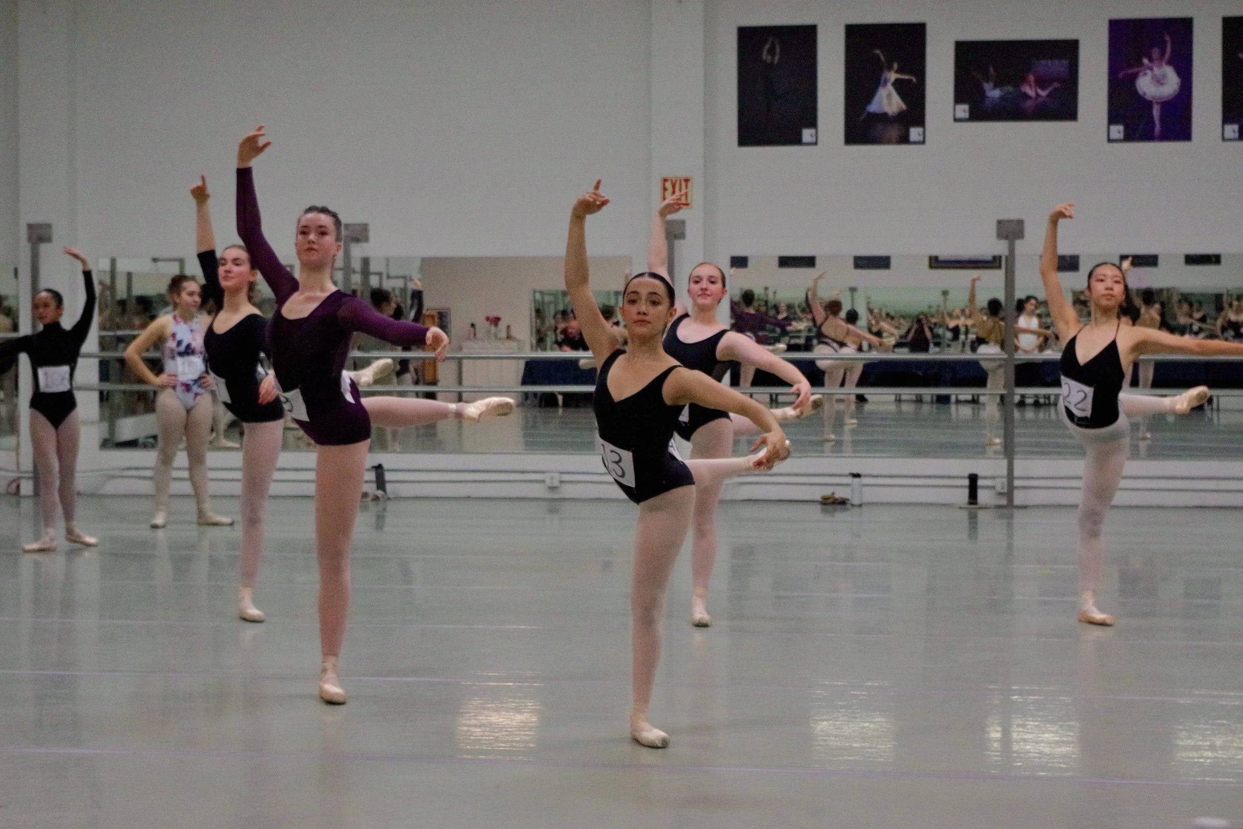 Ballet dancers practicing in a dance studio, performing a ballet pose with arms raised and one leg extended to the side, in front of mirrors and ballet posters.