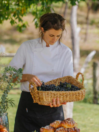 Woman in chef's coat holding a basket of grapes outdoors
