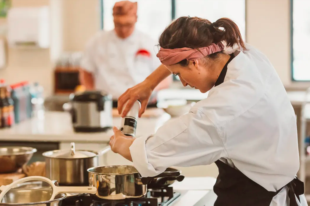 Woman wearing a headband and white chef coat seasoning food on a stove in a kitchen with another chef in the background.