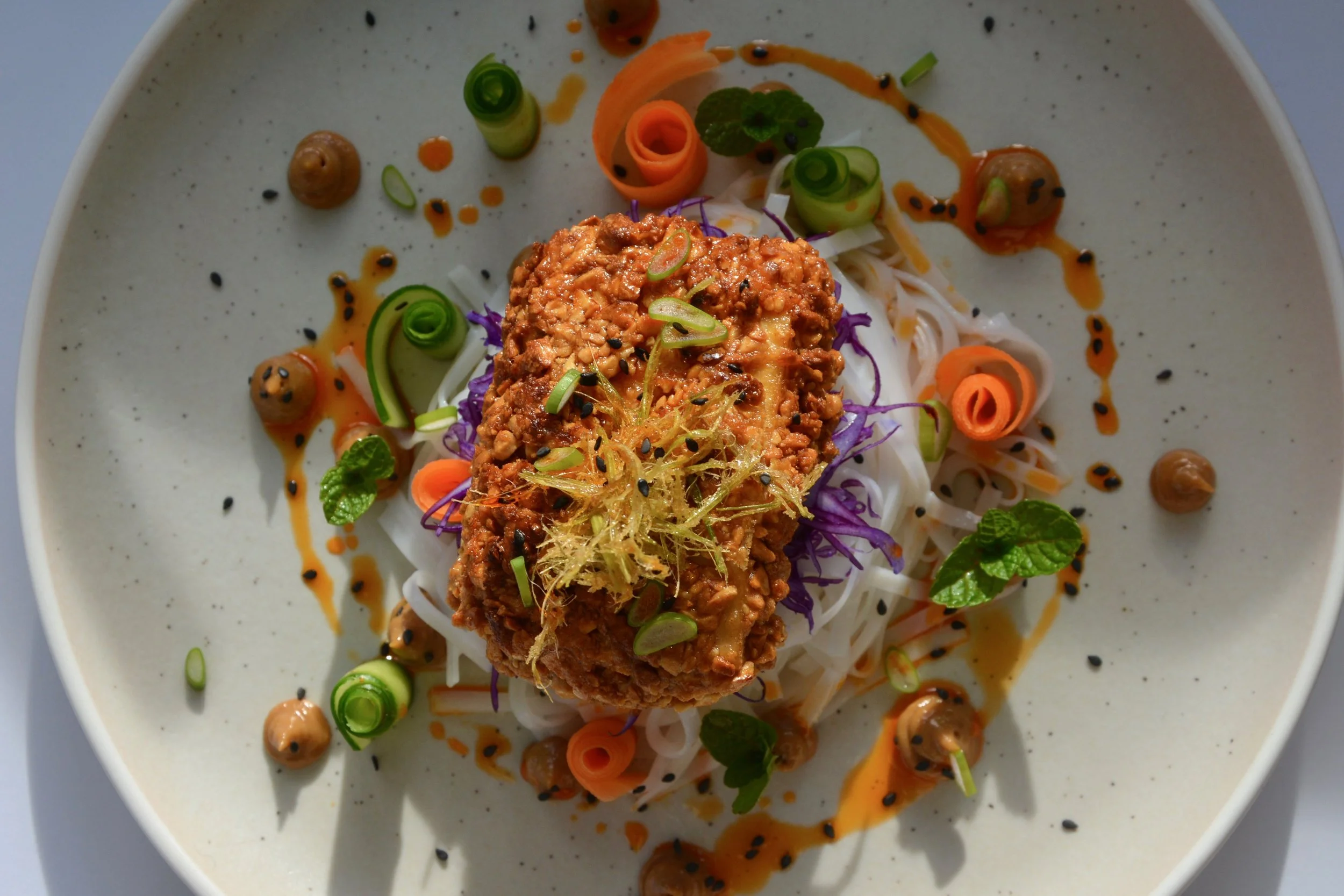 A plate of Asian cuisine featuring a spring roll topped with chopped nuts and garnished with herbs, served over rice noodles with diced vegetables, drizzled with sauce and sprinkled with black sesame seeds.
