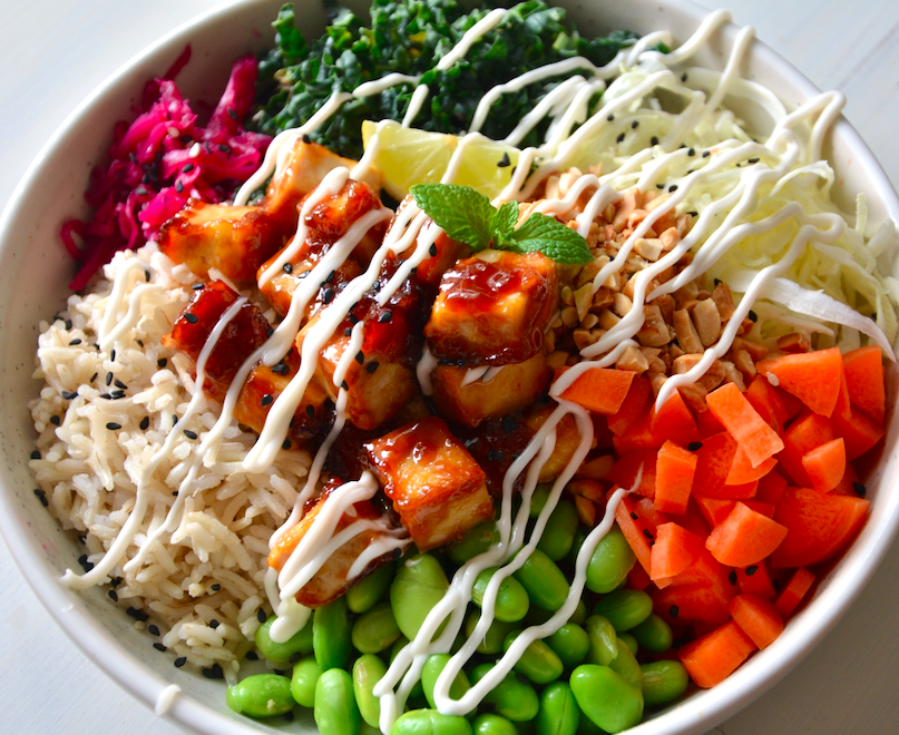 A colorful bowl of rice topped with grilled tofu, cherry tomatoes, shredded cabbage, shredded kale, pickled red cabbage, edamame, and garnished with a lemon wedge and mint leaf, drizzled with white sauce.