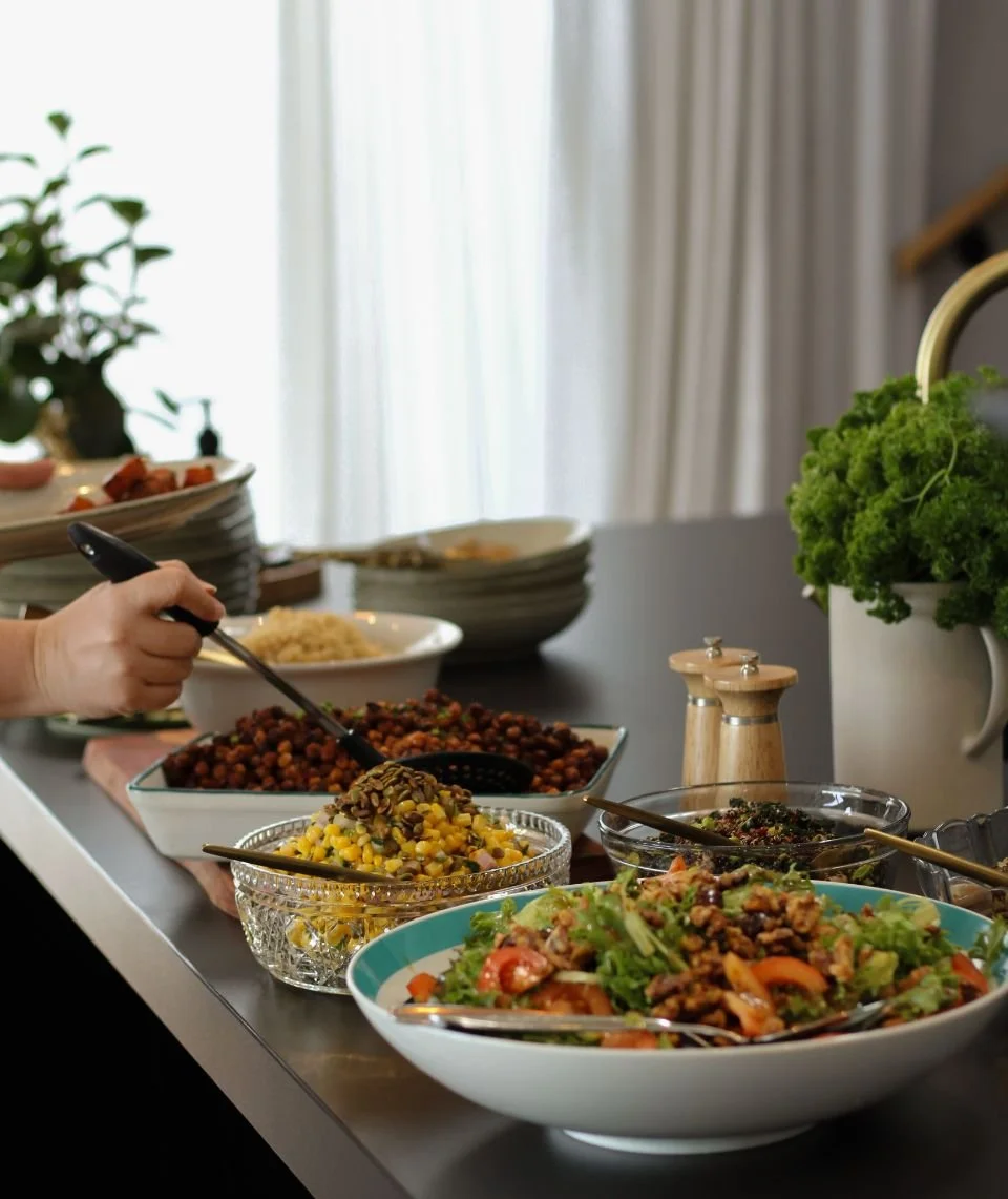 A variety of salads and dishes on a buffet table, including a bowl of mixed greens with vegetables, corn, chickpeas, and other ingredients, with an individual serving salad tongs in use.