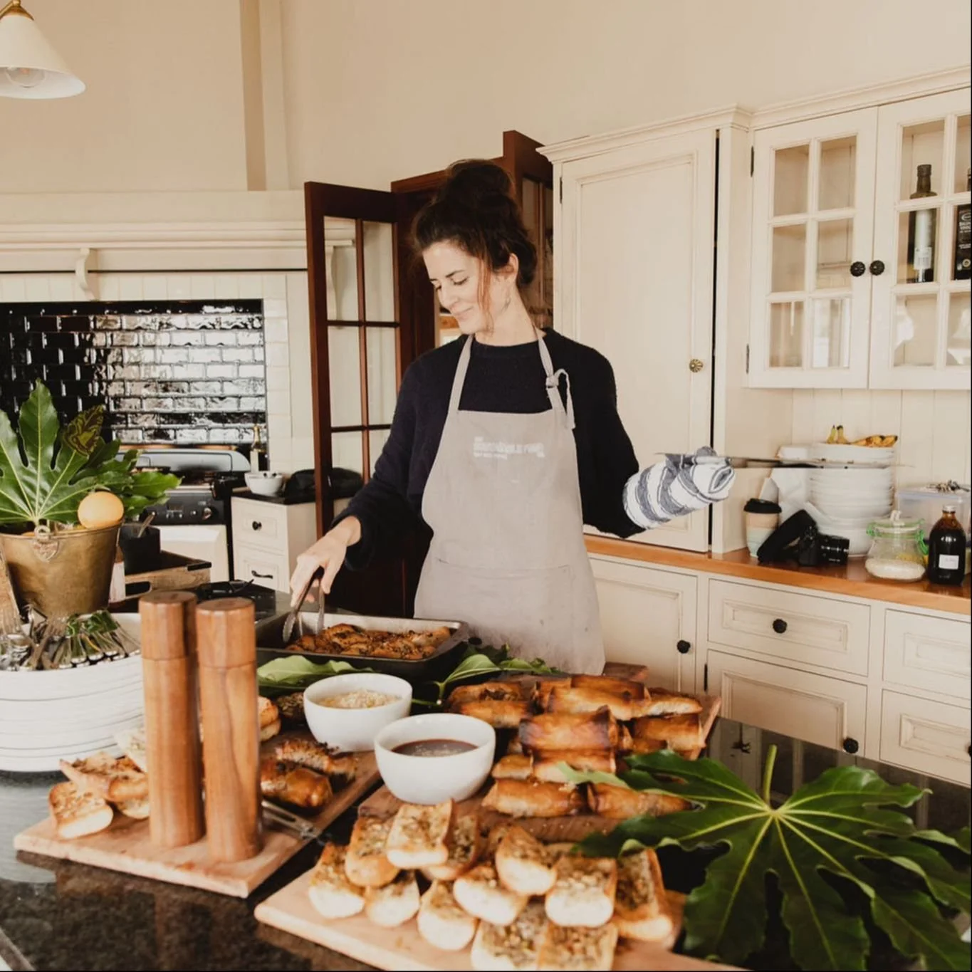 Woman wearing an apron and oven mitts preparing food in a kitchen with a variety of appetizers and snacks on the counter.