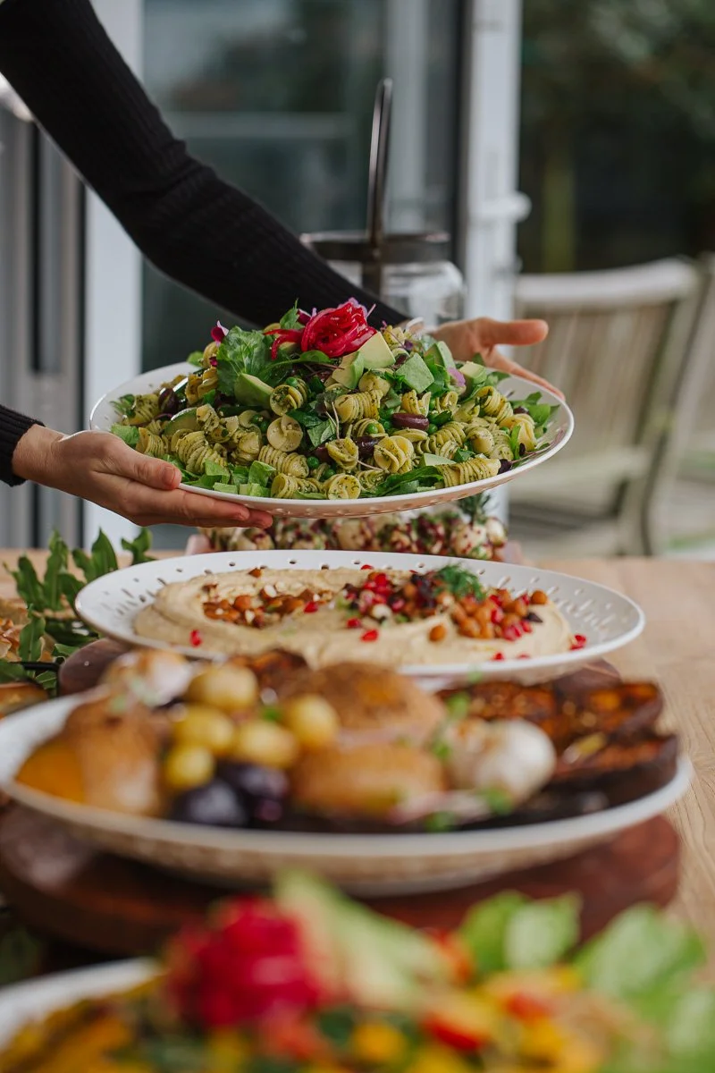 Person serving a pasta salad with green fusilli pasta, vegetables, and herbs onto a plate at a buffet table.