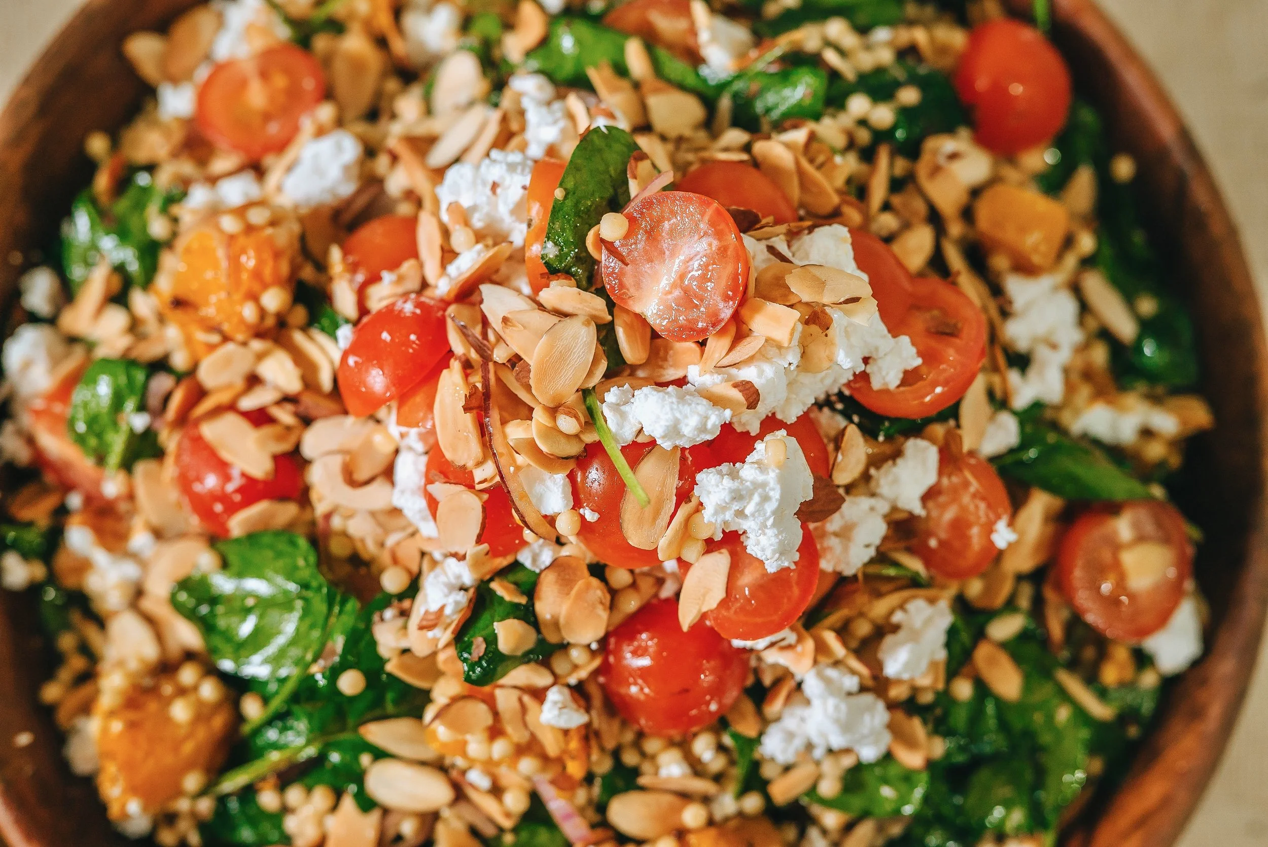 Close-up view of a colorful salad with cherry tomatoes, leafy greens, crumbled cheese, and toasted sliced almonds in a wooden bowl.