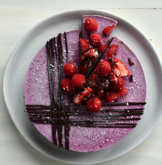 A purple berry-flavored cake decorated with fresh strawberries, raspberries, and chocolate shavings, drizzled with chocolate.
