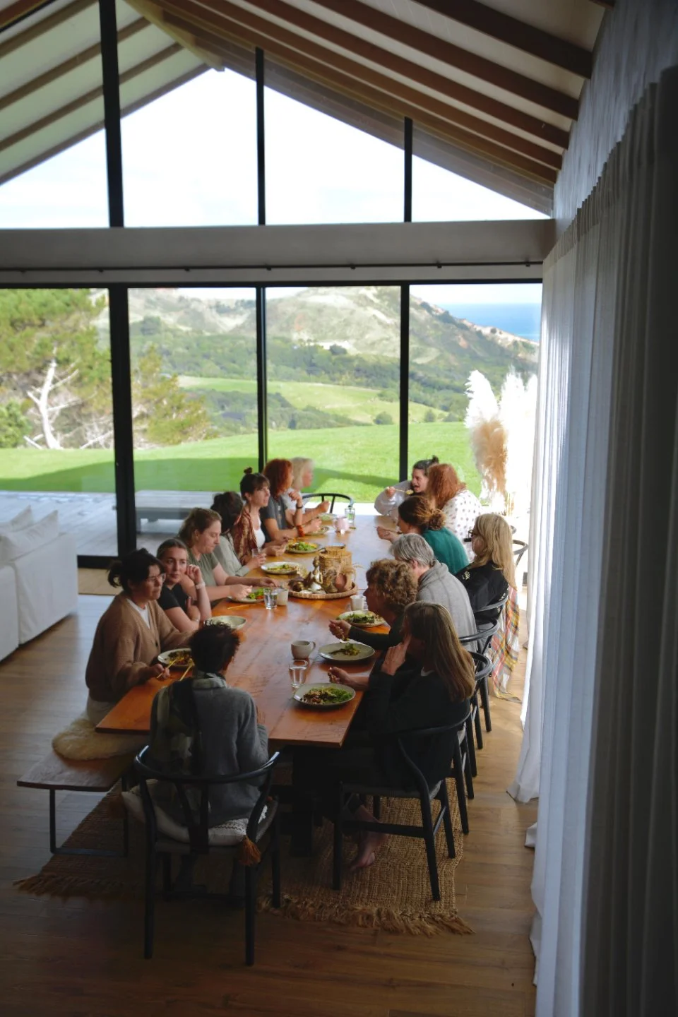 Group of people gathered around a long wooden dining table in a modern house with large glass windows showing a green hillside and mountains in the background, enjoying a meal.