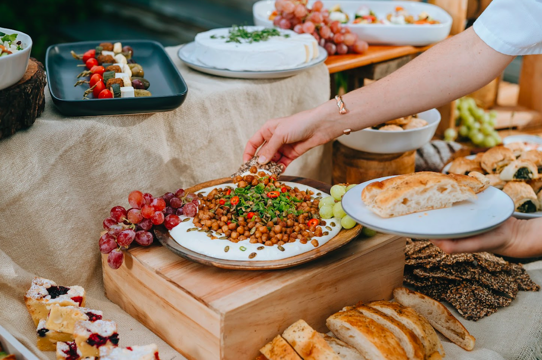 Person serving chickpea salad with bread on a table with grapes and various dishes at a buffet.