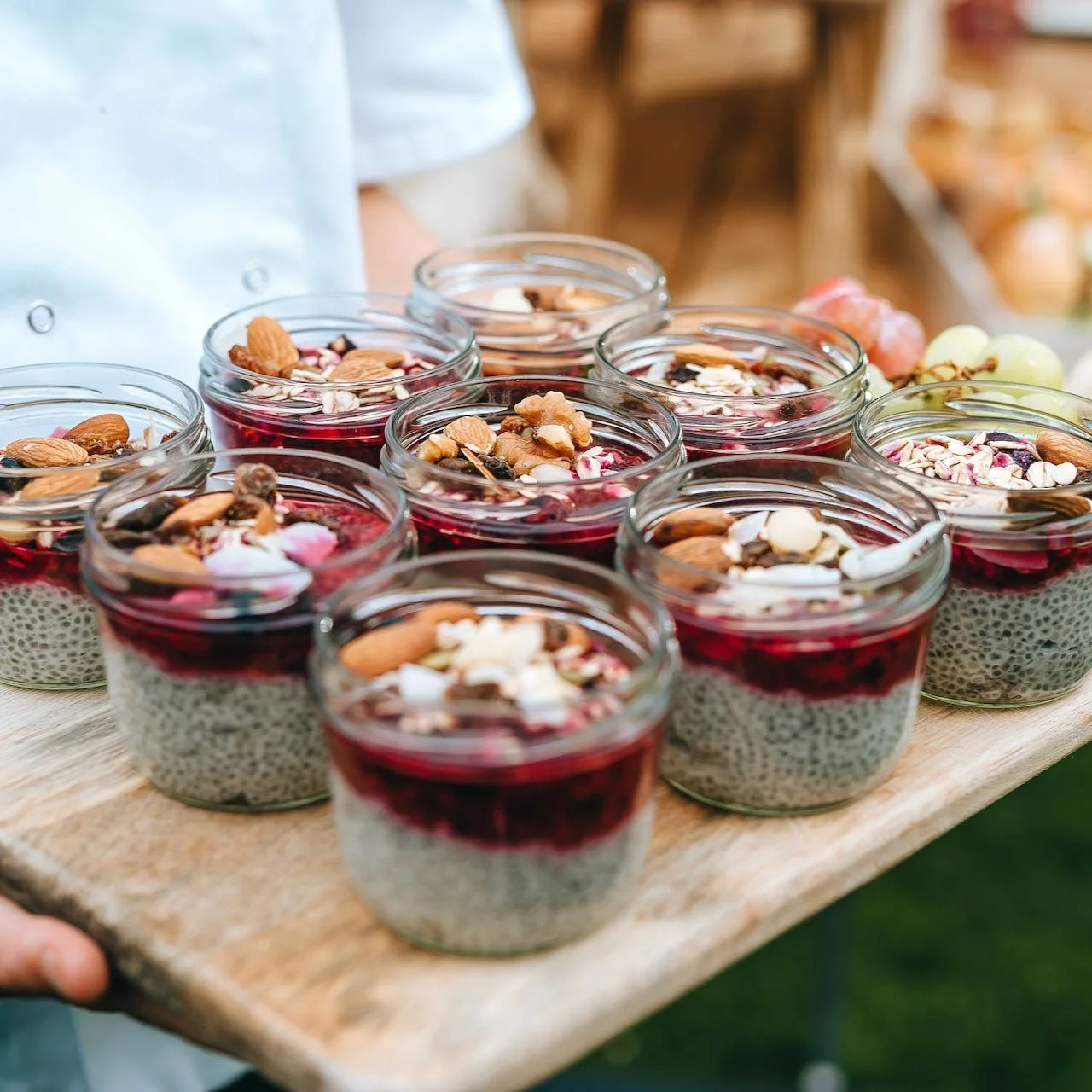 A person holding a wooden tray filled with small glass jars containing chia seed pudding topped with mixed nuts and berries.