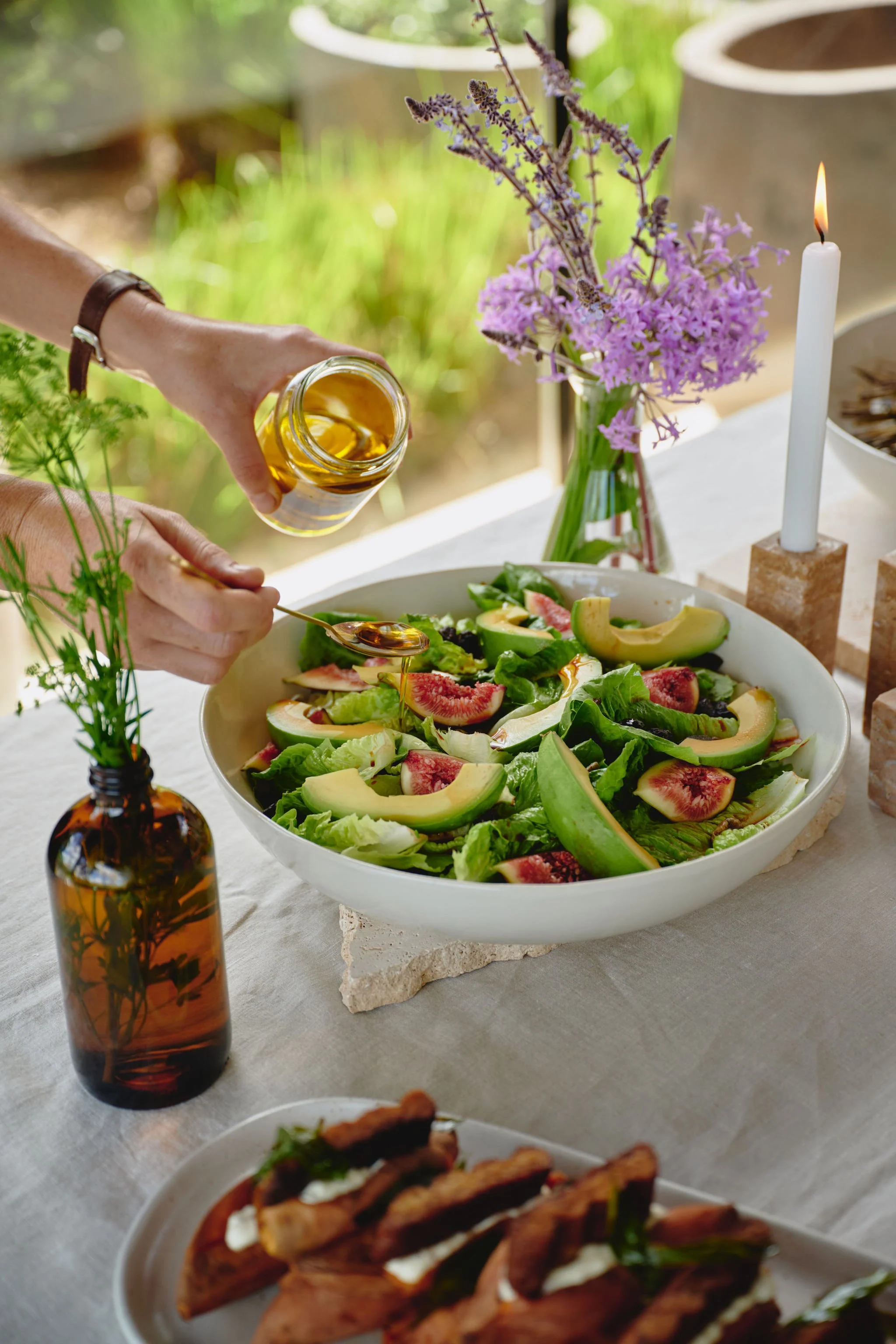 A person pouring olive oil on a fresh salad with figs and greens, with a lit candle and a vase of lavender flowers on the table.