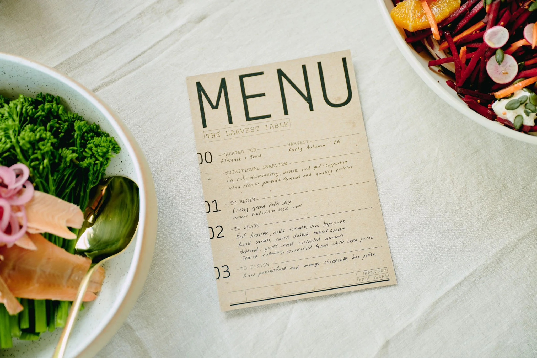 A restaurant menu titled "The Harvest Table" on a light-colored table with two bowls of salad, one containing greens with pink slices and other vegetables, partially visible, and another containing red and orange vegetables, radishes, and dressing.