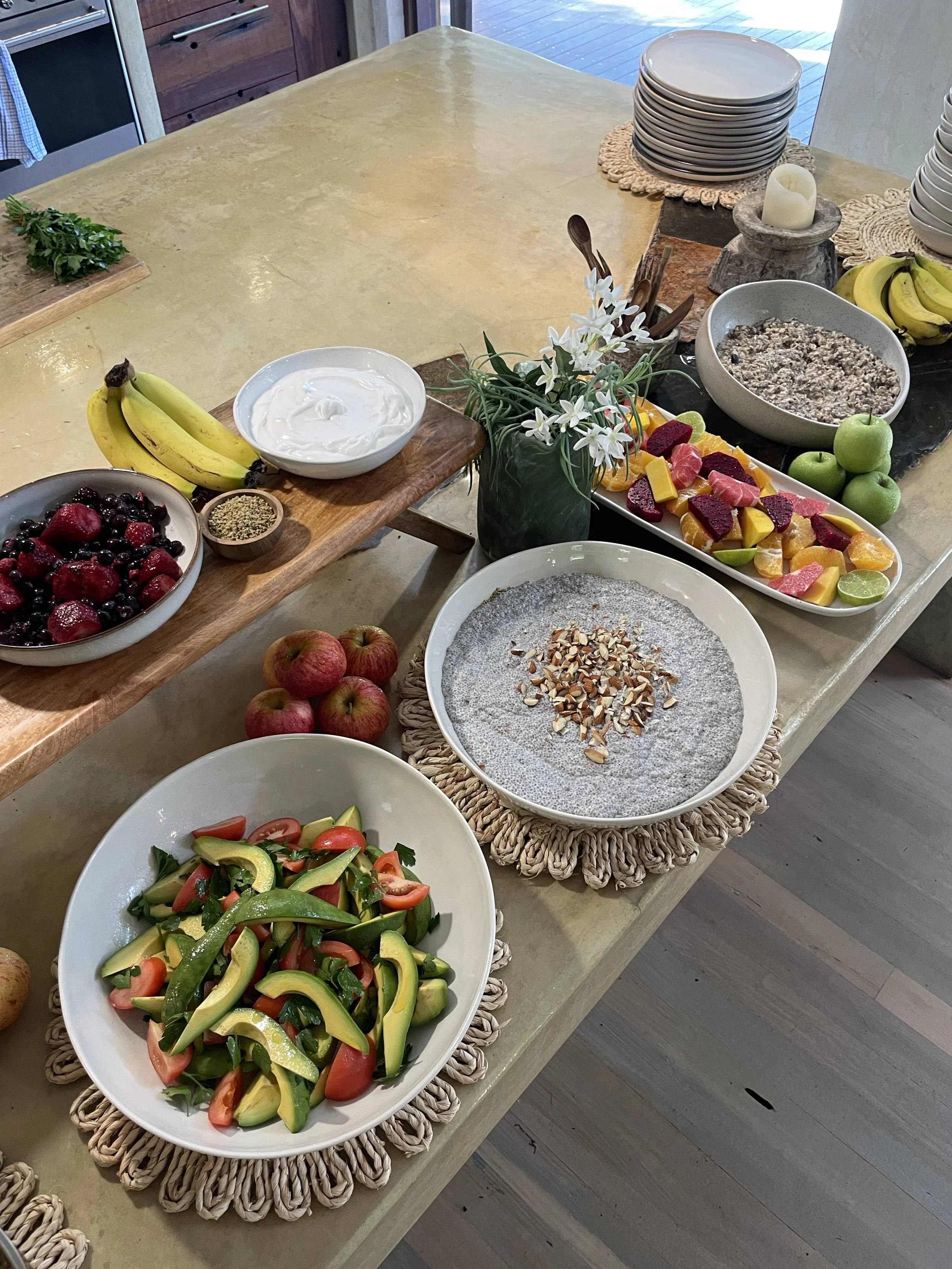 A dining table with various bowls of fruit and salad, including bananas, strawberries, blueberries, oranges, grapefruit, apples, chopped vegetables, and granola, on woven placemats.