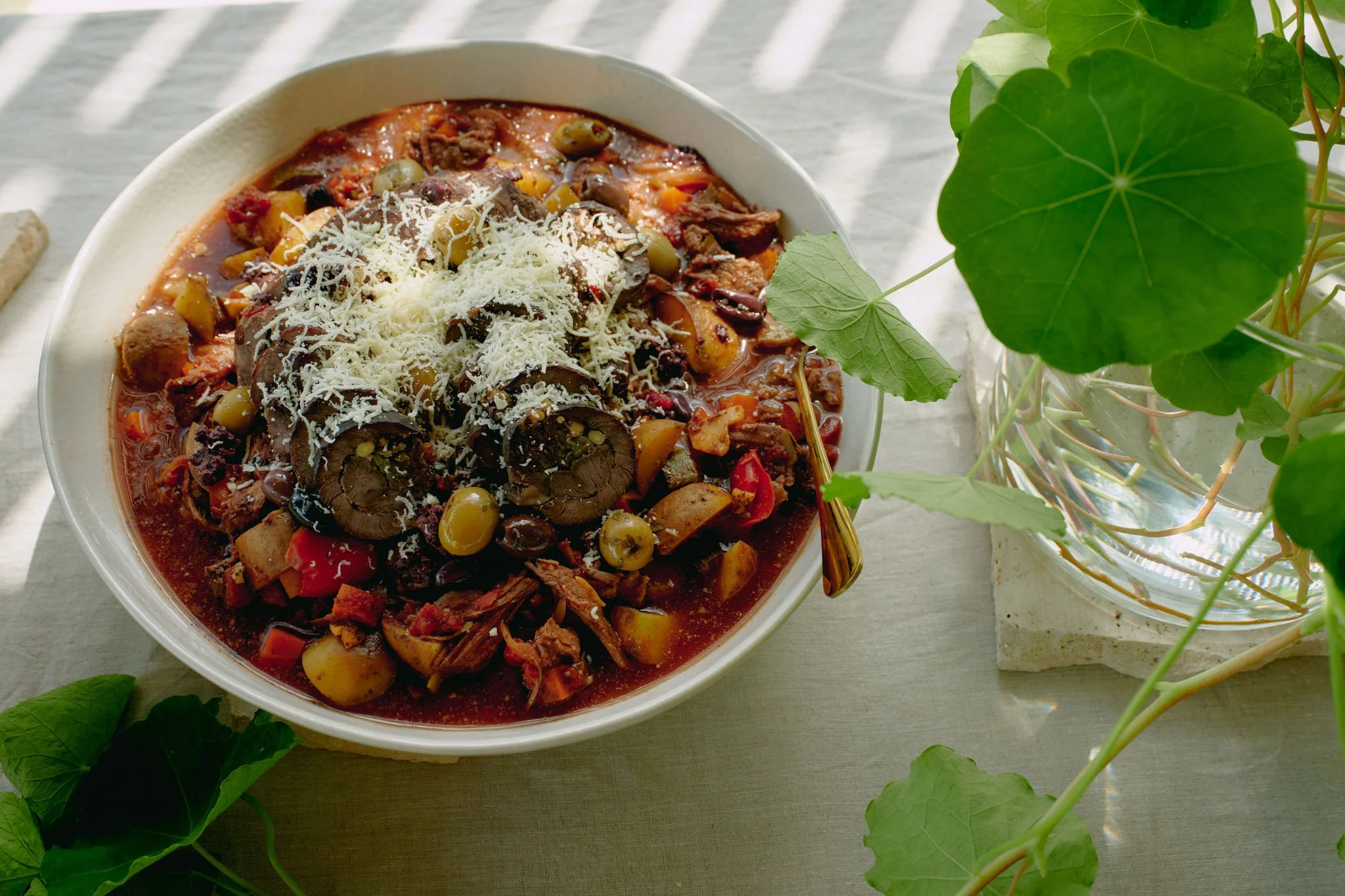 A bowl of chili with beans, shredded cheese, and meat on a white surface, with green leaves nearby.