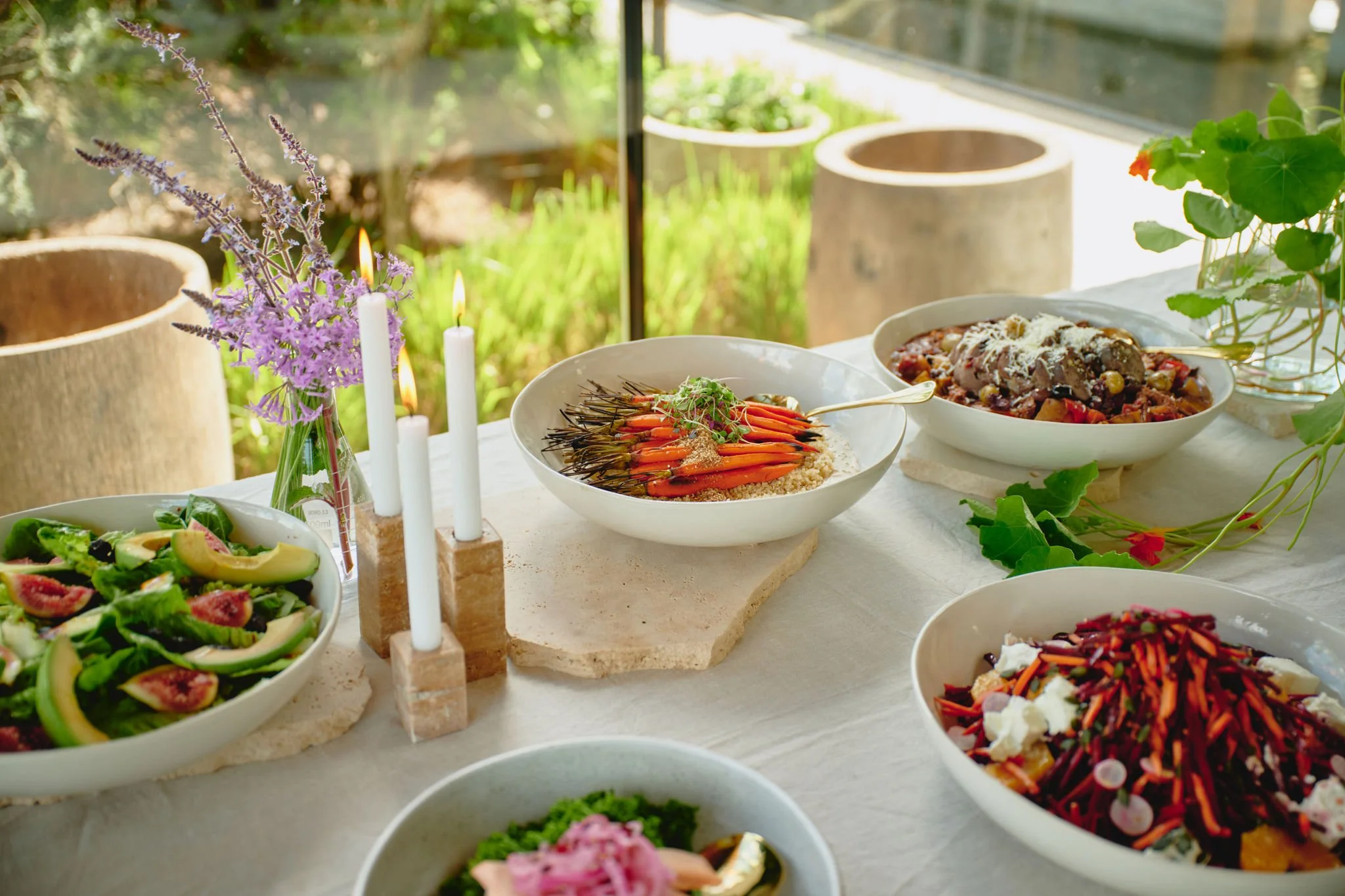 Arrangement of colorful salads and dishes on a white table with candles, flowers, and potted plants in the background, suggesting a garden or outdoor dining setting.
