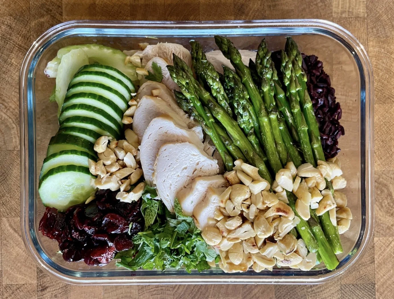 A glass container filled with sliced cucumbers, roasted asparagus, sliced turkey breast, chopped nuts, mixed greens, and black rice, all arranged on a wooden surface.