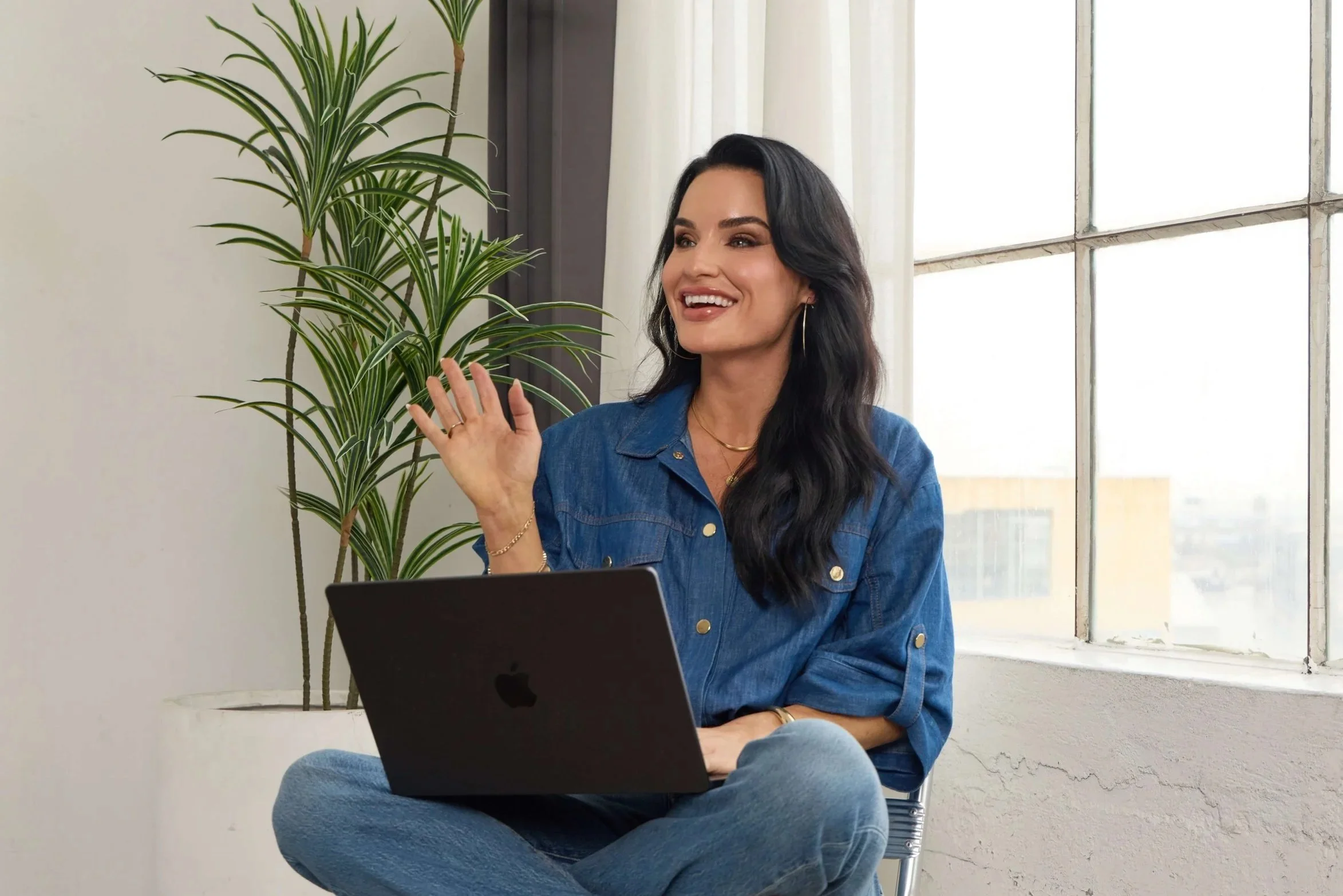 A woman with long black hair, smiling and waving, sitting on a chair with a black laptop on her lap, wearing a blue denim shirt and light blue jeans, in a bright room with large window and a potted plant nearby.