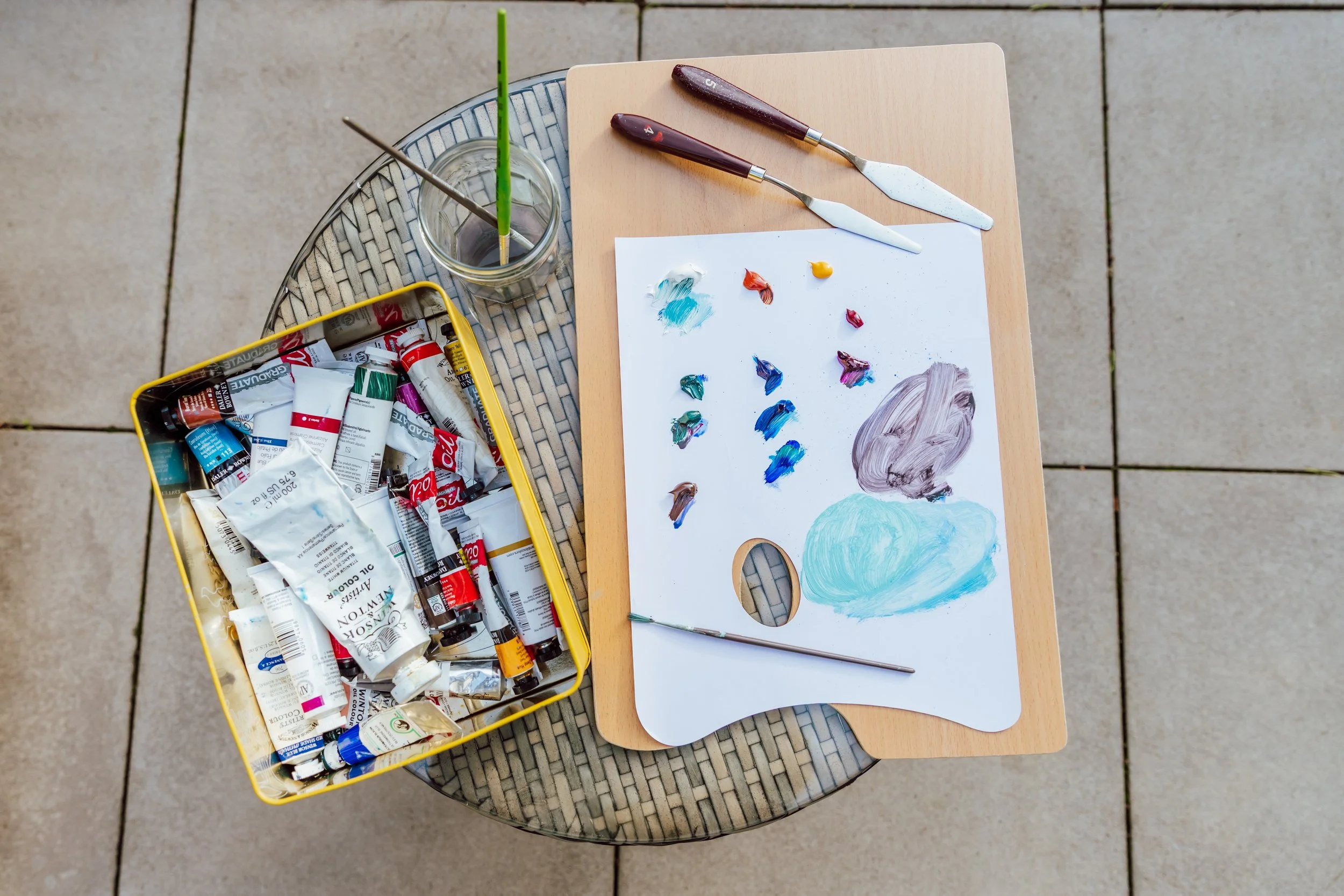 An art easel with a palette of mixed oil paints, two paintbrushes, and a container of paint tubes on a small round table outside.