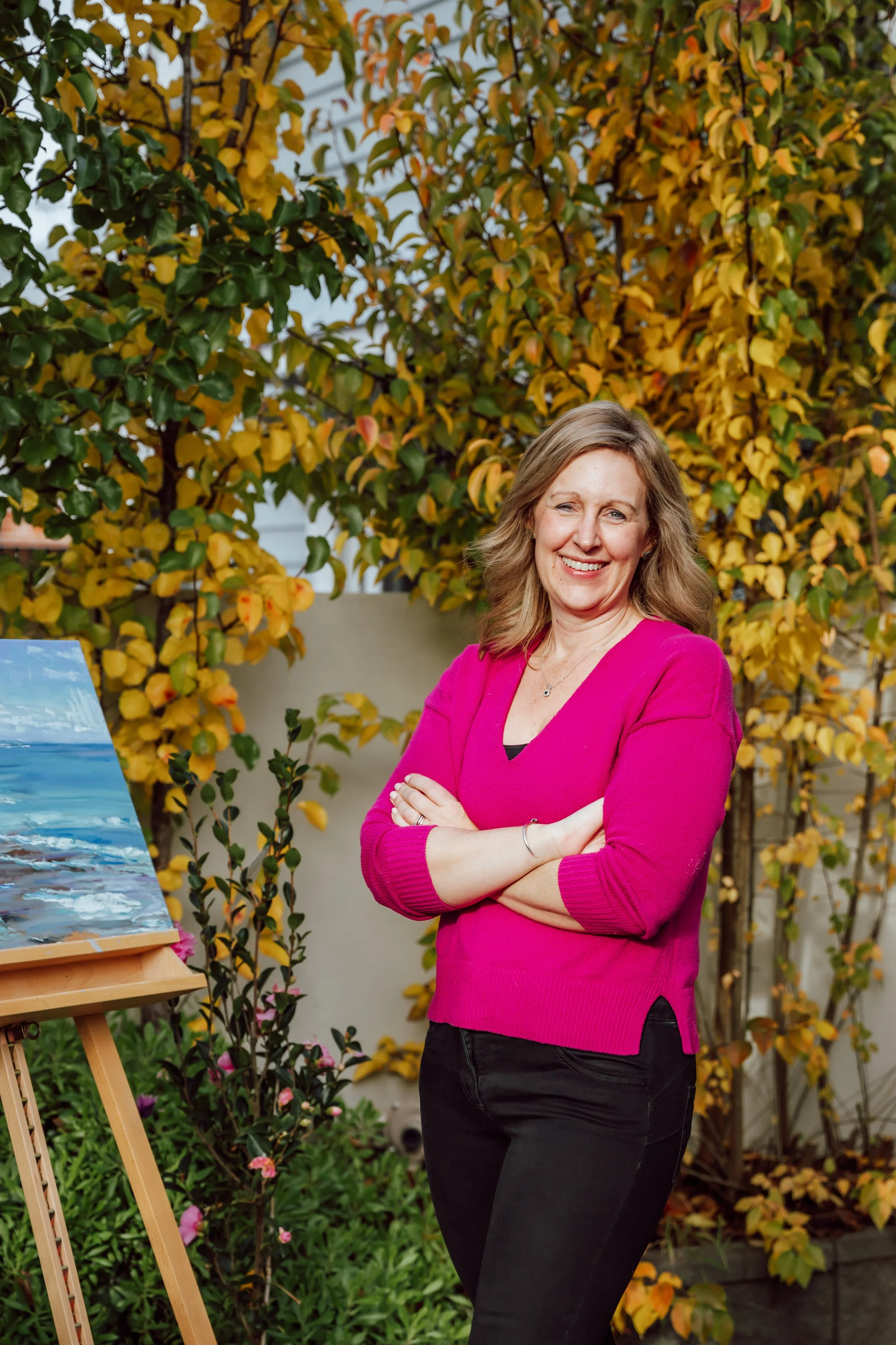 A woman with wavy blonde hair, wearing a bright pink sweater and black pants, smiling with her arms crossed, standing outdoors next to an easel with a landscape painting of a beach scene, surrounded by green and yellow foliage.