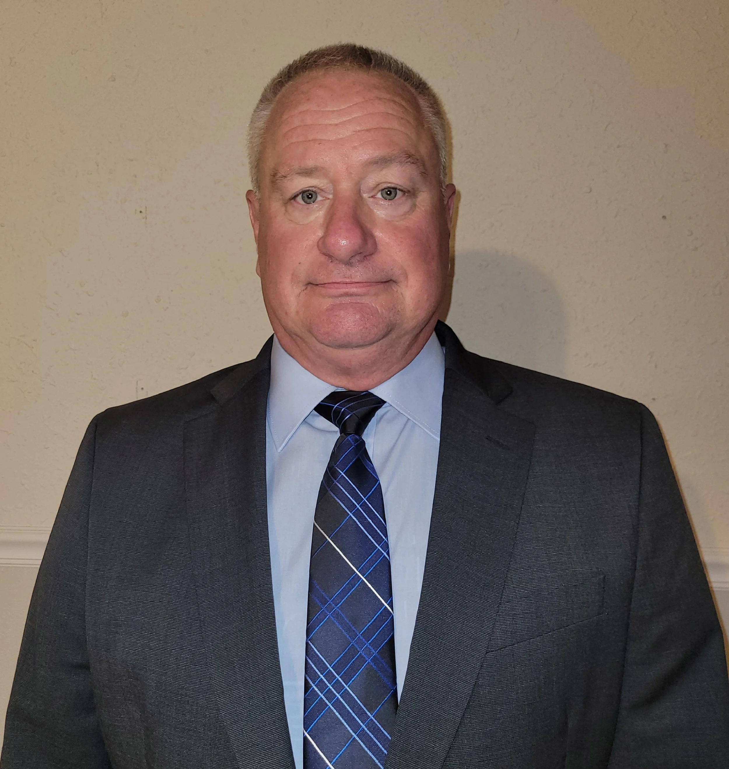 A middle-aged man with light hair and a slight smile, wearing a dark suit, light blue shirt, and a blue plaid tie, standing against a beige wall.