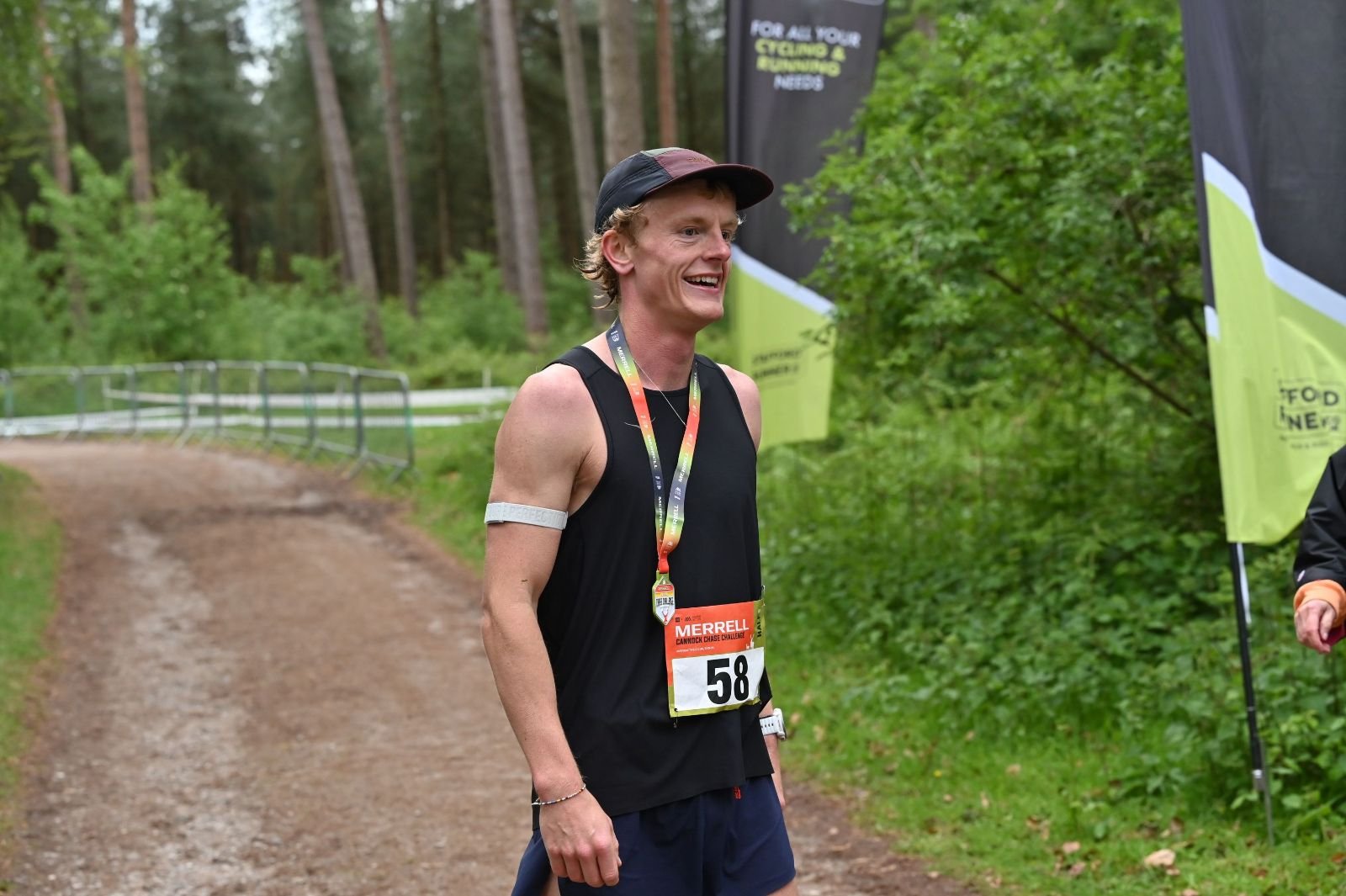 A male runner in a black sleeveless shirt and navy shorts standing on a dirt trail with green trees in the background, smiling after completing a race, wearing a race bib with the number 58 and a medal around his neck.