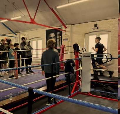 Group of people inside a boxing gym with a boxing ring, trainers, and workout equipment.