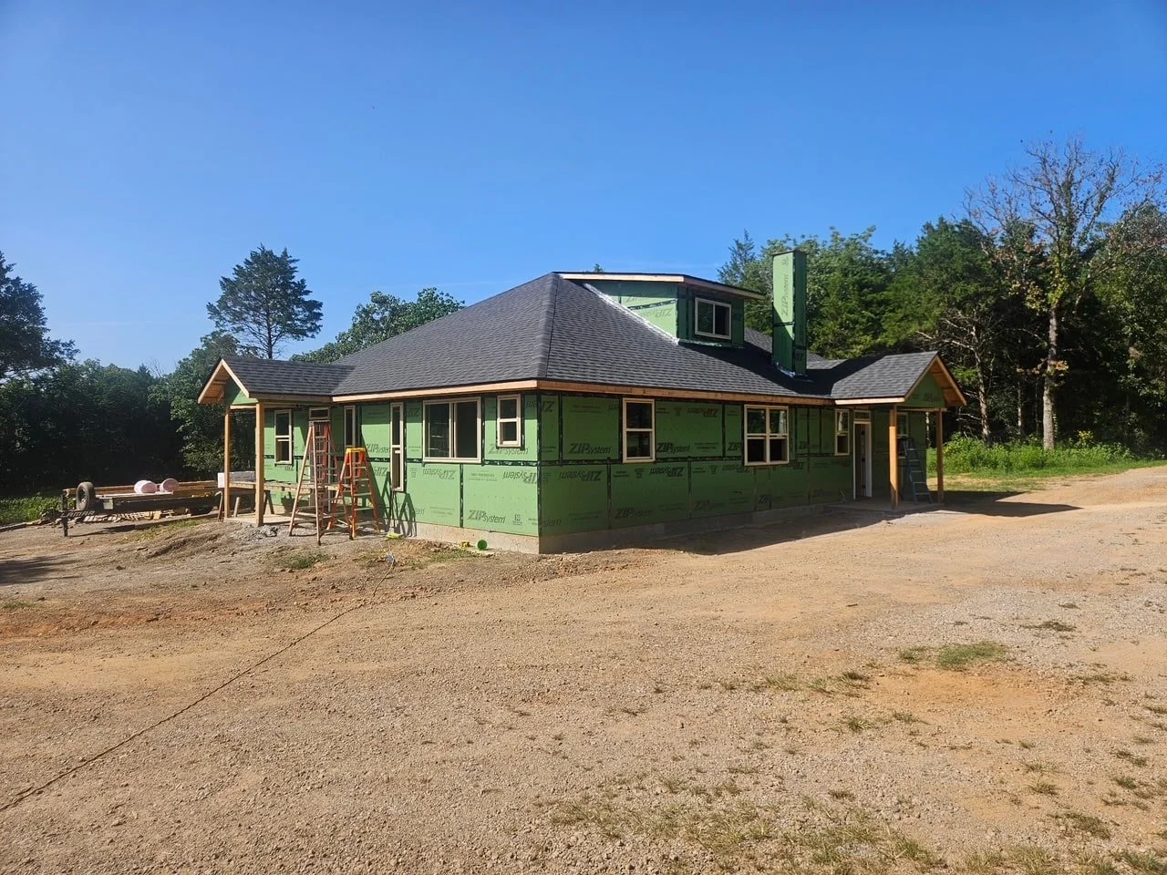 A house under construction with green sheathing and a dark gray roof, surrounded by dirt and trees in the background.