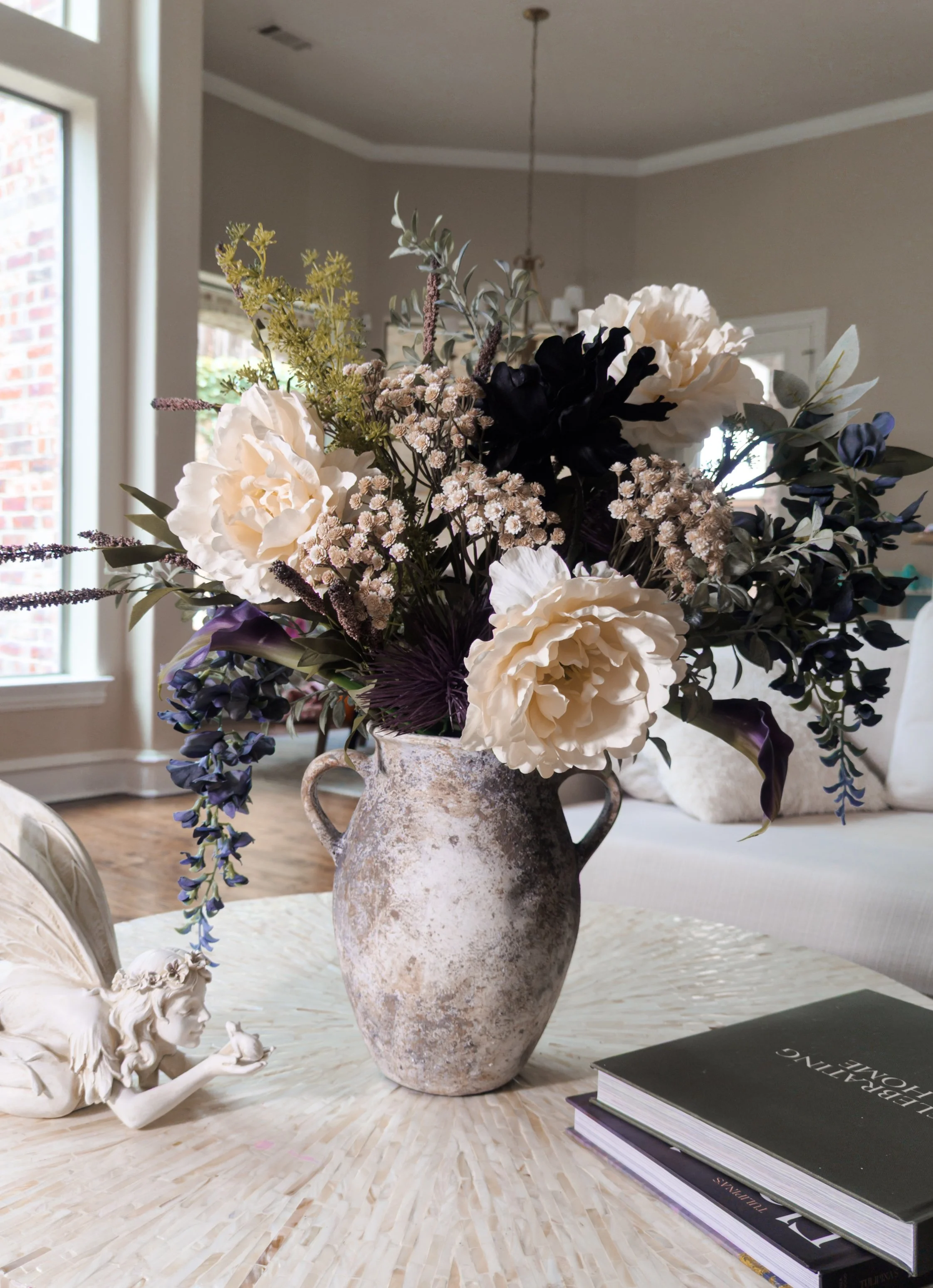A rustic ceramic vase filled with a large arrangement of white, purple, and dark green artificial flowers on a woven table. Next to the vase, a small angel figurine with a bow and wing is blowing a kiss. Two books are stacked nearby, one titled "LEAD