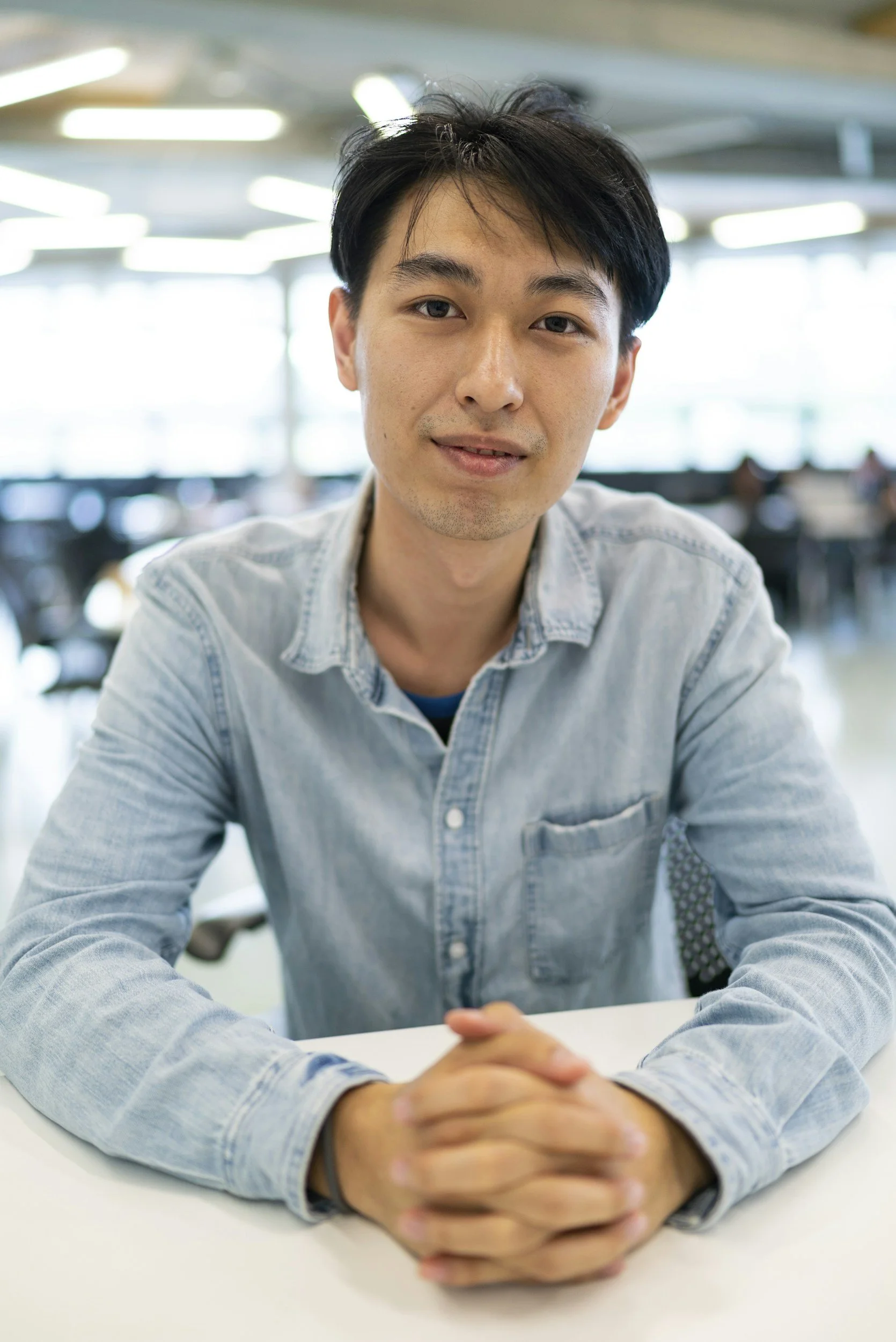 A young man with black hair, wearing a light denim shirt, sitting at a table in a bright, modern office space with large windows.