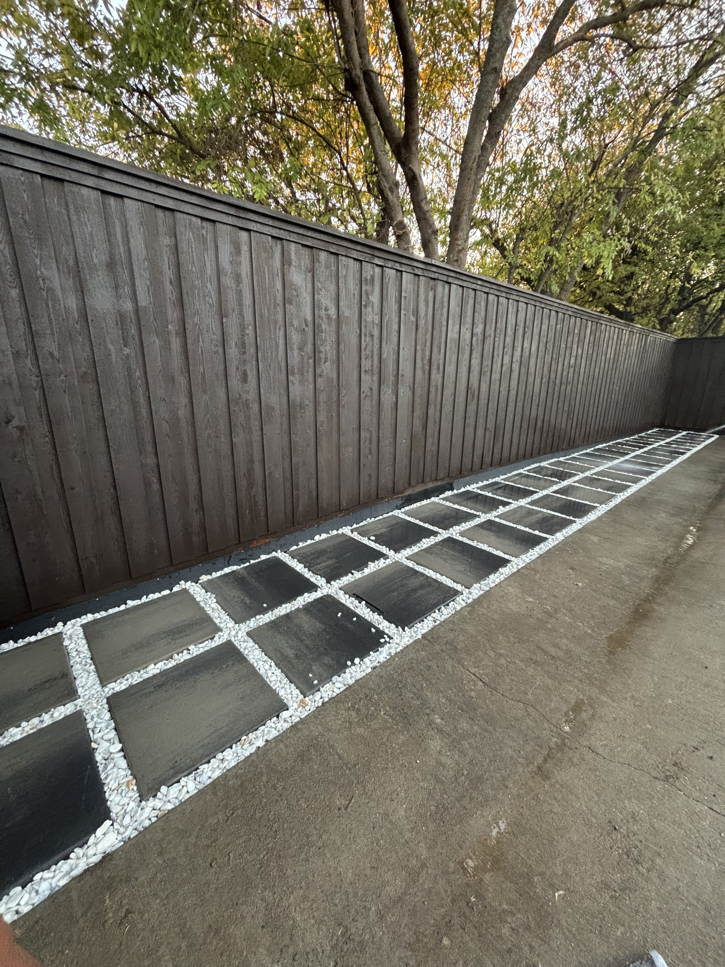 Sidewalk with black tiles bordered by white pebbles, next to a wooden fence with trees in the background.