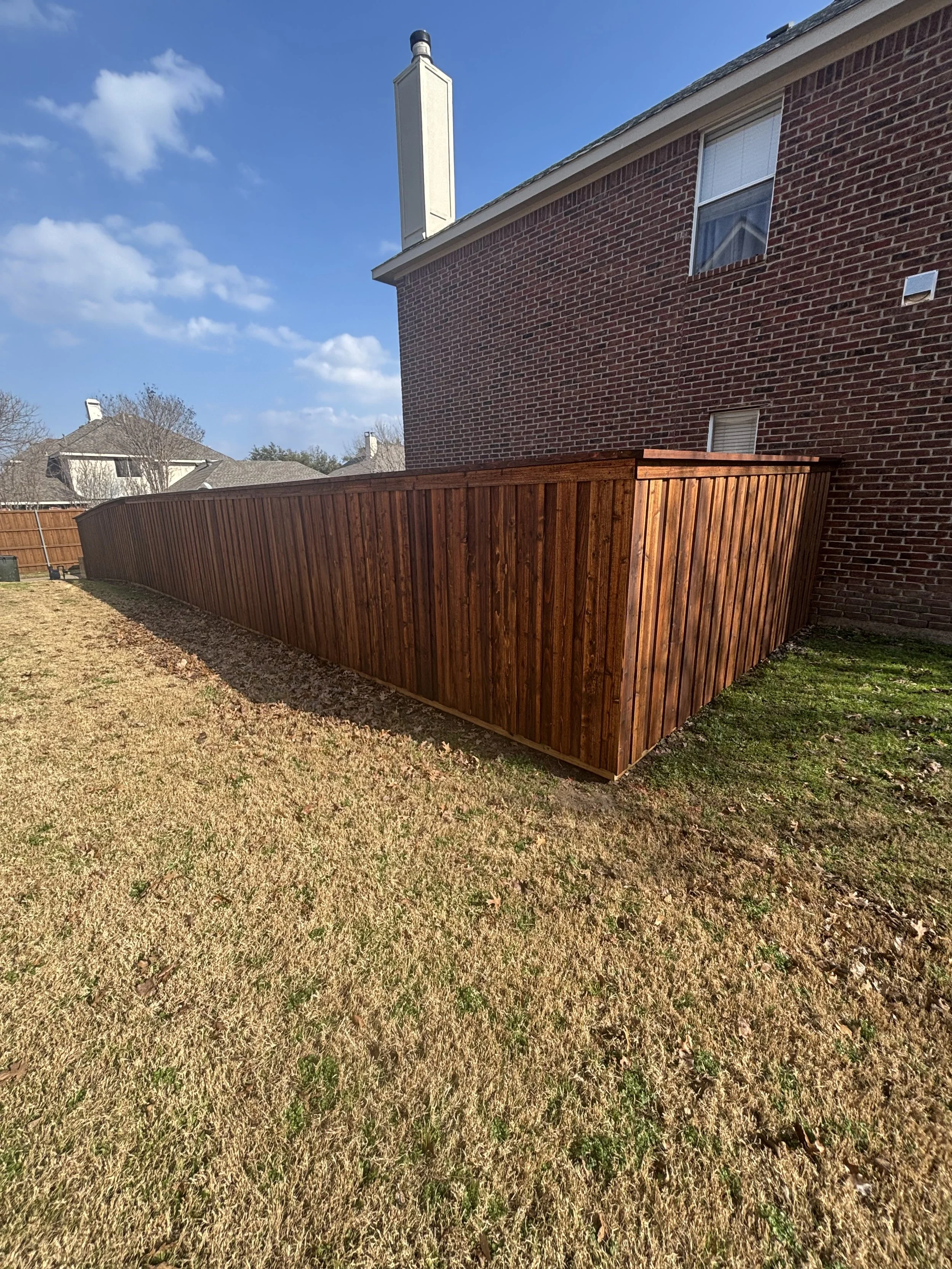 A backyard with a tall wooden privacy fence adjacent to a brick house, with a chimney on top, under a blue sky with some clouds.