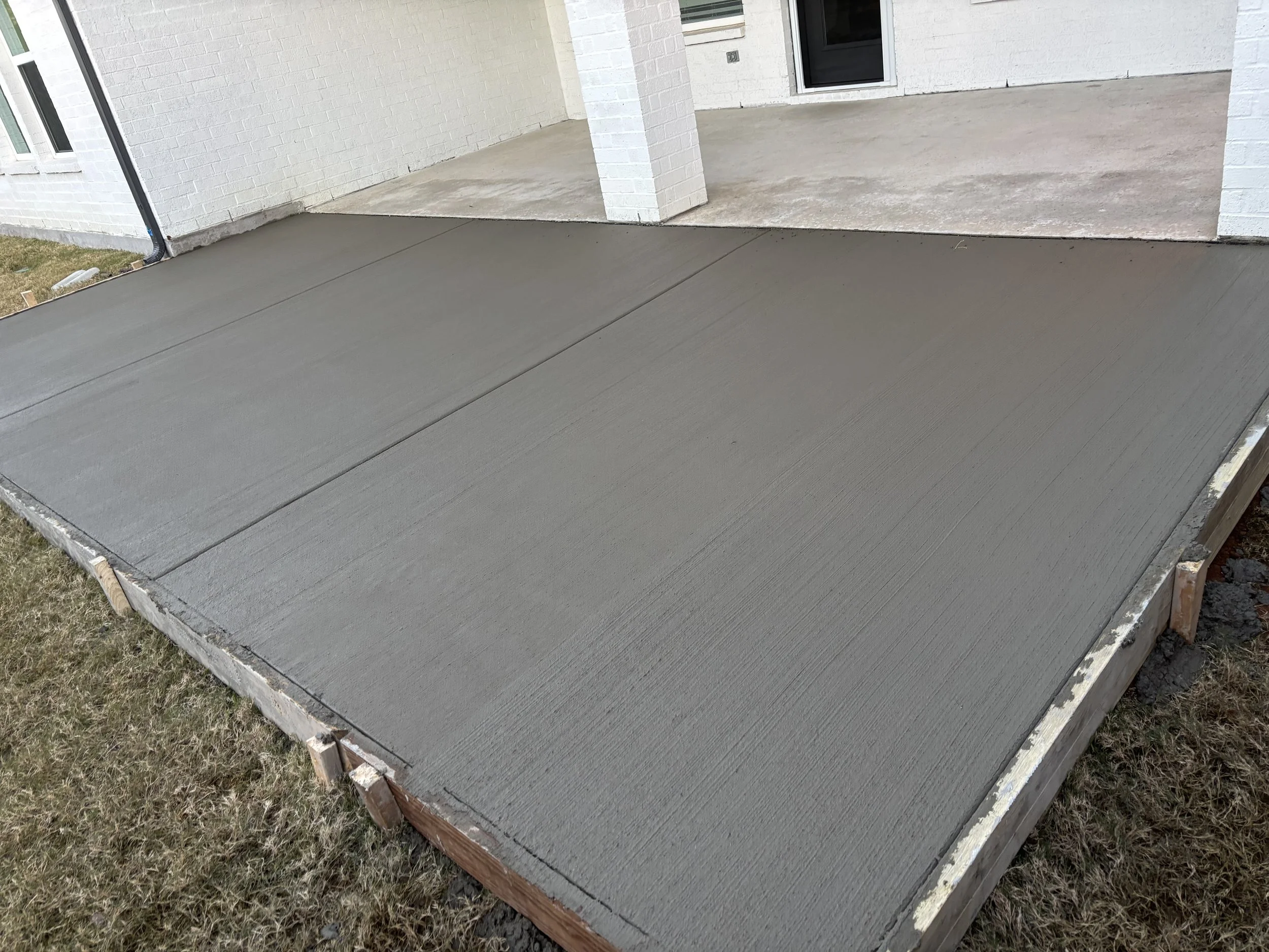 Freshly poured concrete slab on a residential porch with white brick exterior walls and grass surrounding the area.