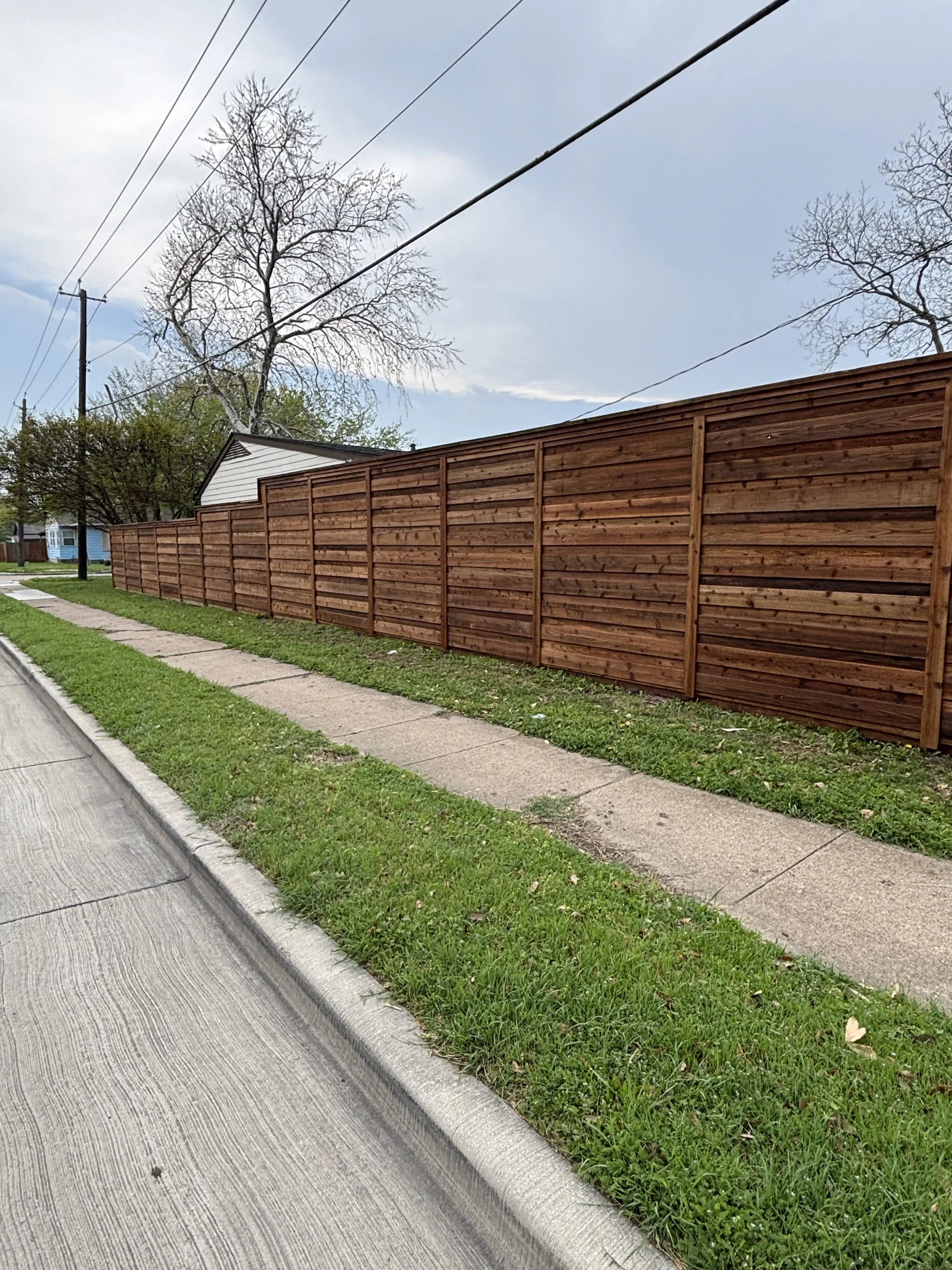 A wooden privacy fence along a sidewalk with a grassy strip, under partly cloudy sky, with some trees and a house in the background.