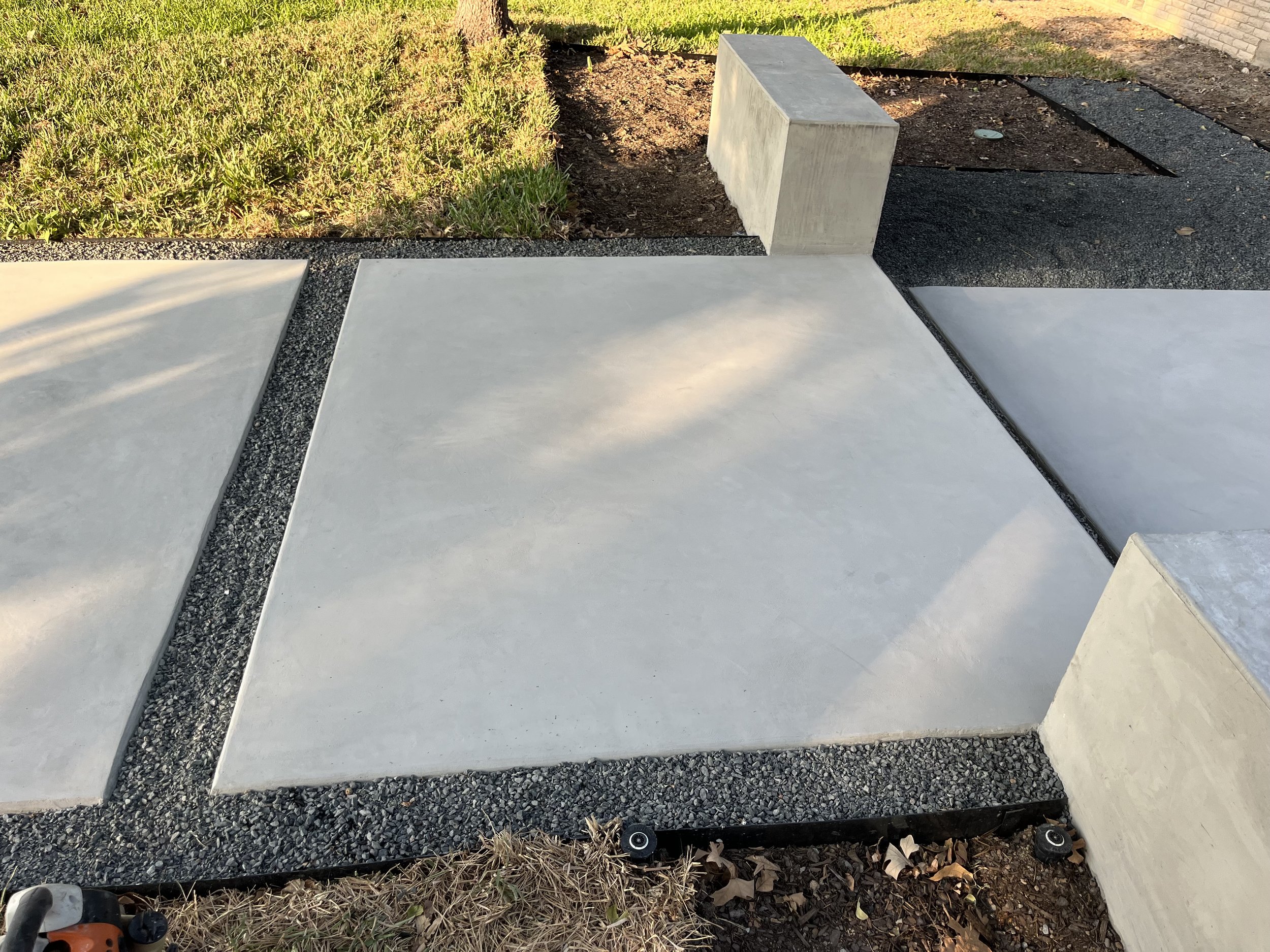 Concrete slabs with gravel between them, part of a walkway or patio, with a small patch of grass and planting area at the top, and a brick wall on the right.