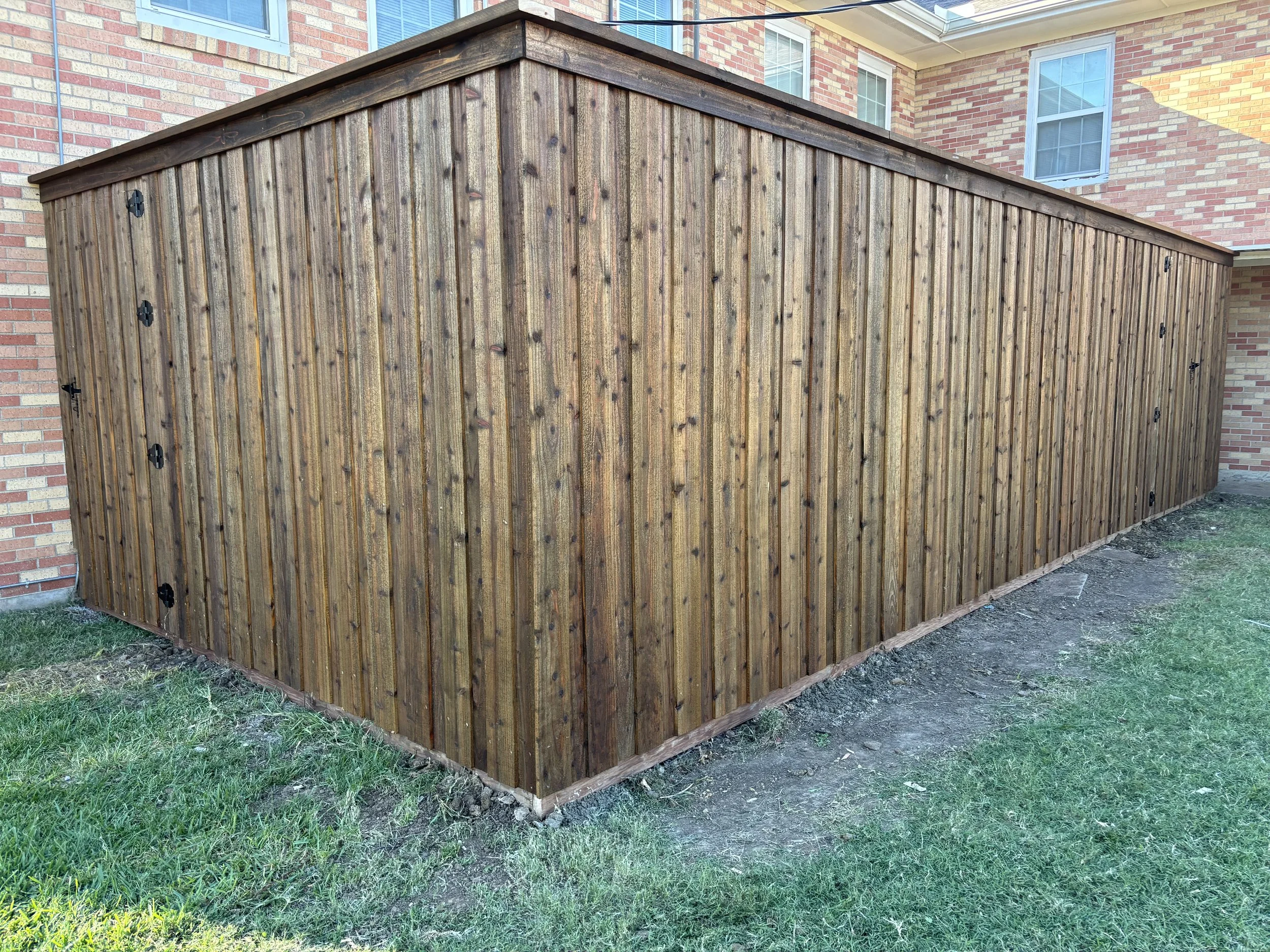 Wooden shed with vertical planks attached to brick house exterior, located on a grassy yard with some dirt patches.