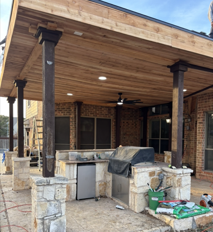 Under construction outdoor covered patio with stone columns, ceiling fan, and partially finished wooden ceiling, featuring built-in kitchen area with countertop, small fridge, and grill, surrounded by construction tools and materials.